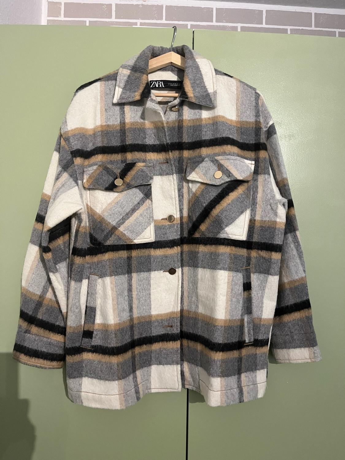 White, grey, black and beige checked overshirt on a wooden hanger against a green background. ZARA text visible on the inside label.