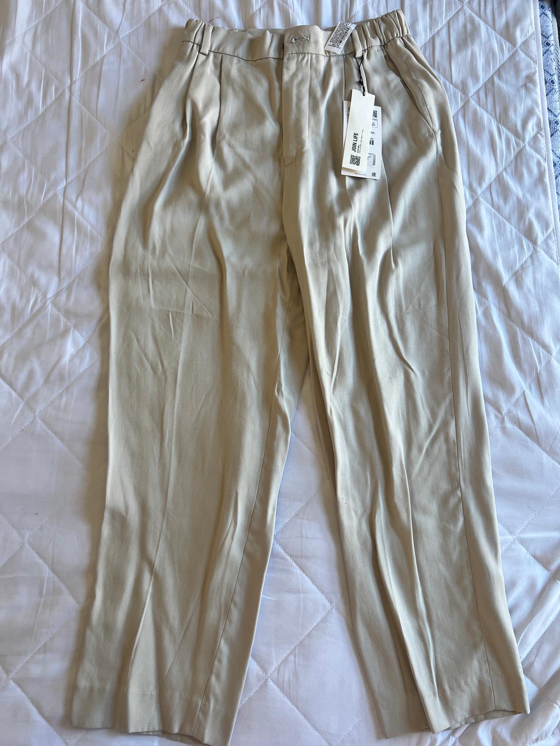 Lightweight beige trousers with an elasticated waist and hang tag, on a white quilted bedspread.