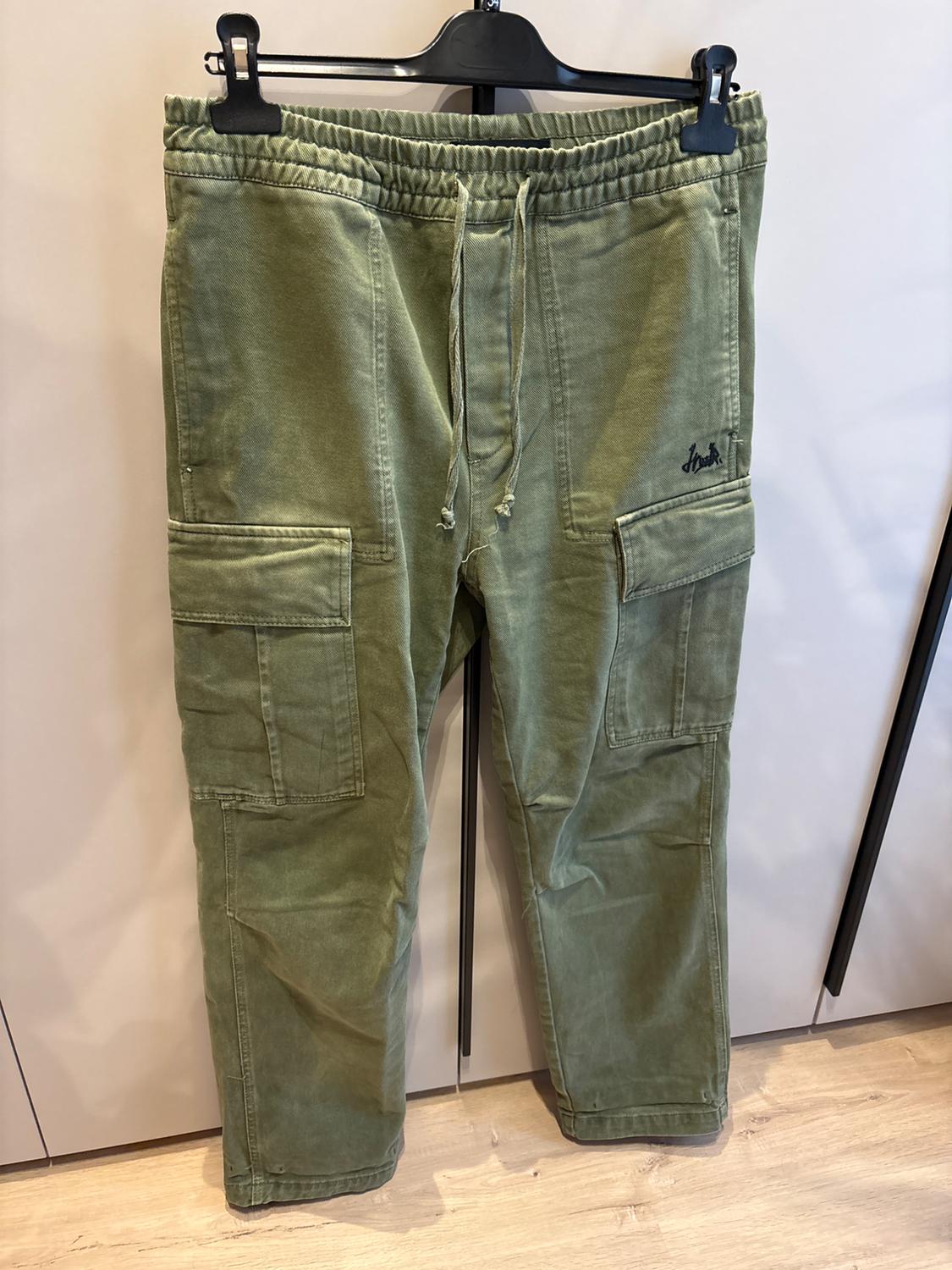 Green cargo trousers hanging on a black hanger, with large pockets and an elasticated drawstring waist. Wooden floor visible.