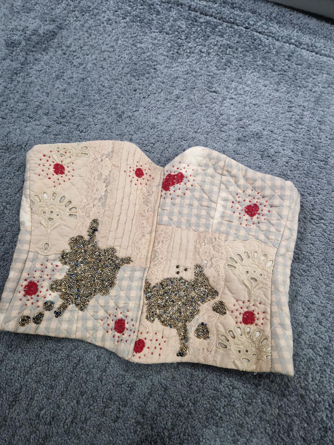 Padded corset in beige and blue tones with lace and bead embroidery, on a grey rug. Design with floral and animal details.