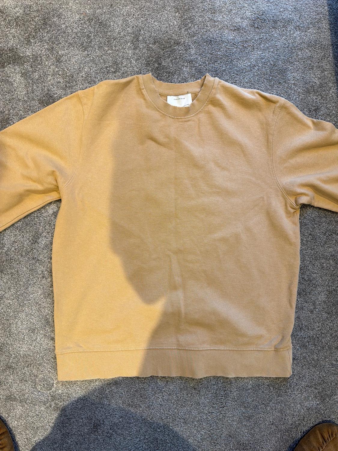 Camel-coloured crew-neck sweatshirt with long sleeves, laid out on a grey rug with shadows cast across the garment.