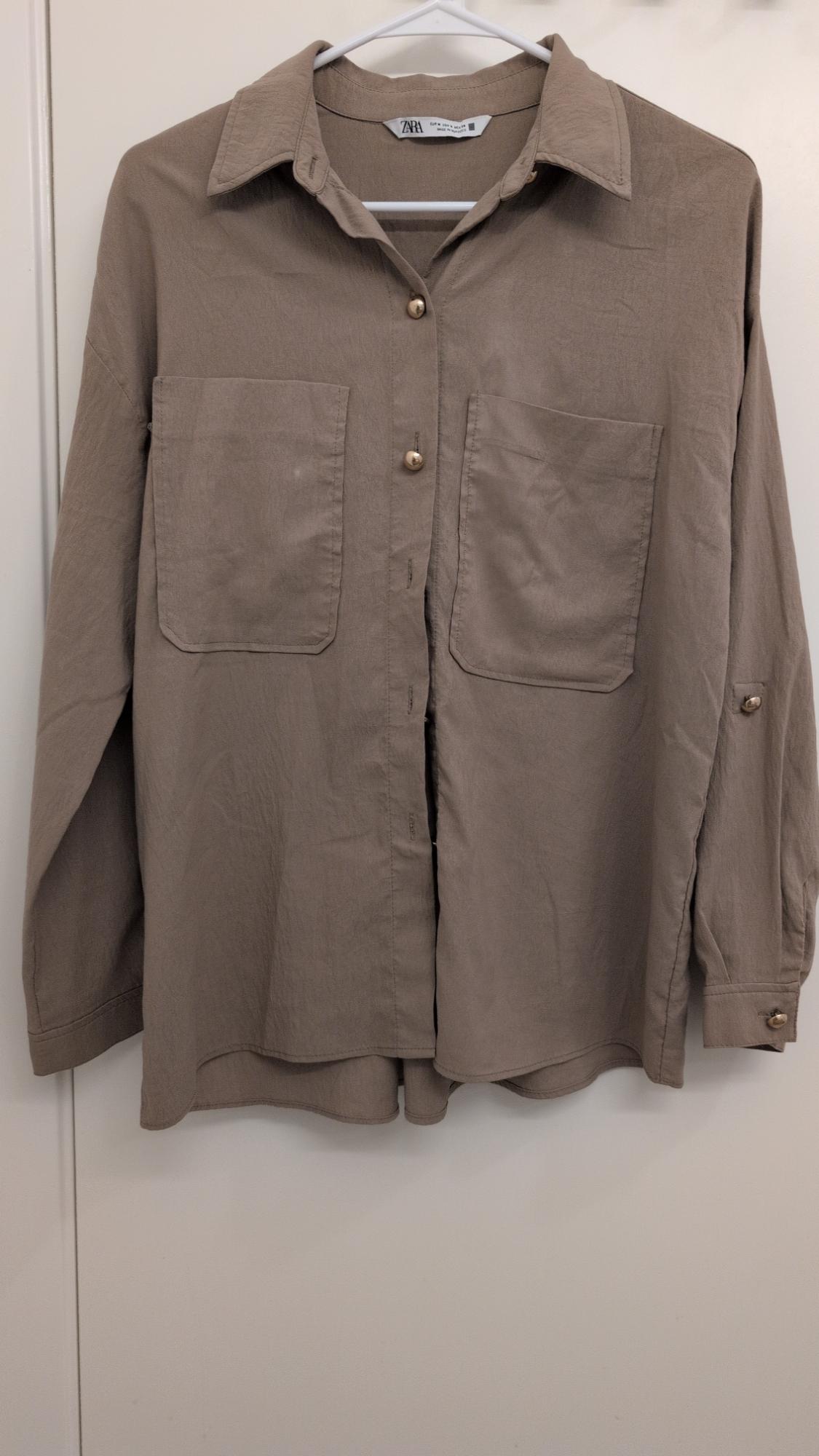 Beige long-sleeved shirt with two front pockets and gold buttons, hanging on a white hanger. Visible label: Zara.