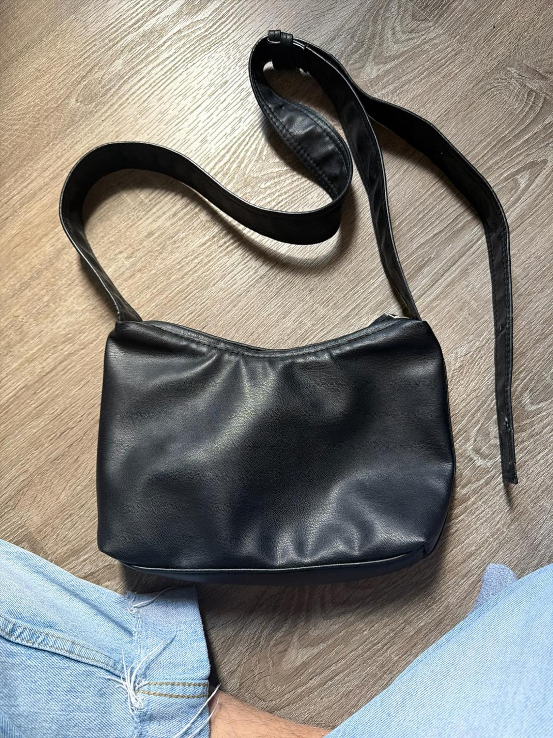 Black bag with a strap on a wooden floor. At the bottom,
legs wearing ripped blue jeans. Casual style.