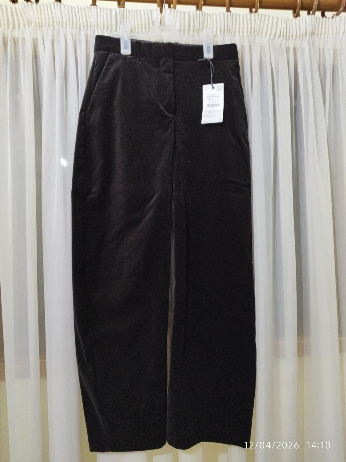 Black trousers hanging on a hanger in front of white curtains. Label visible. Date in image: 12/04/2026 14:10.