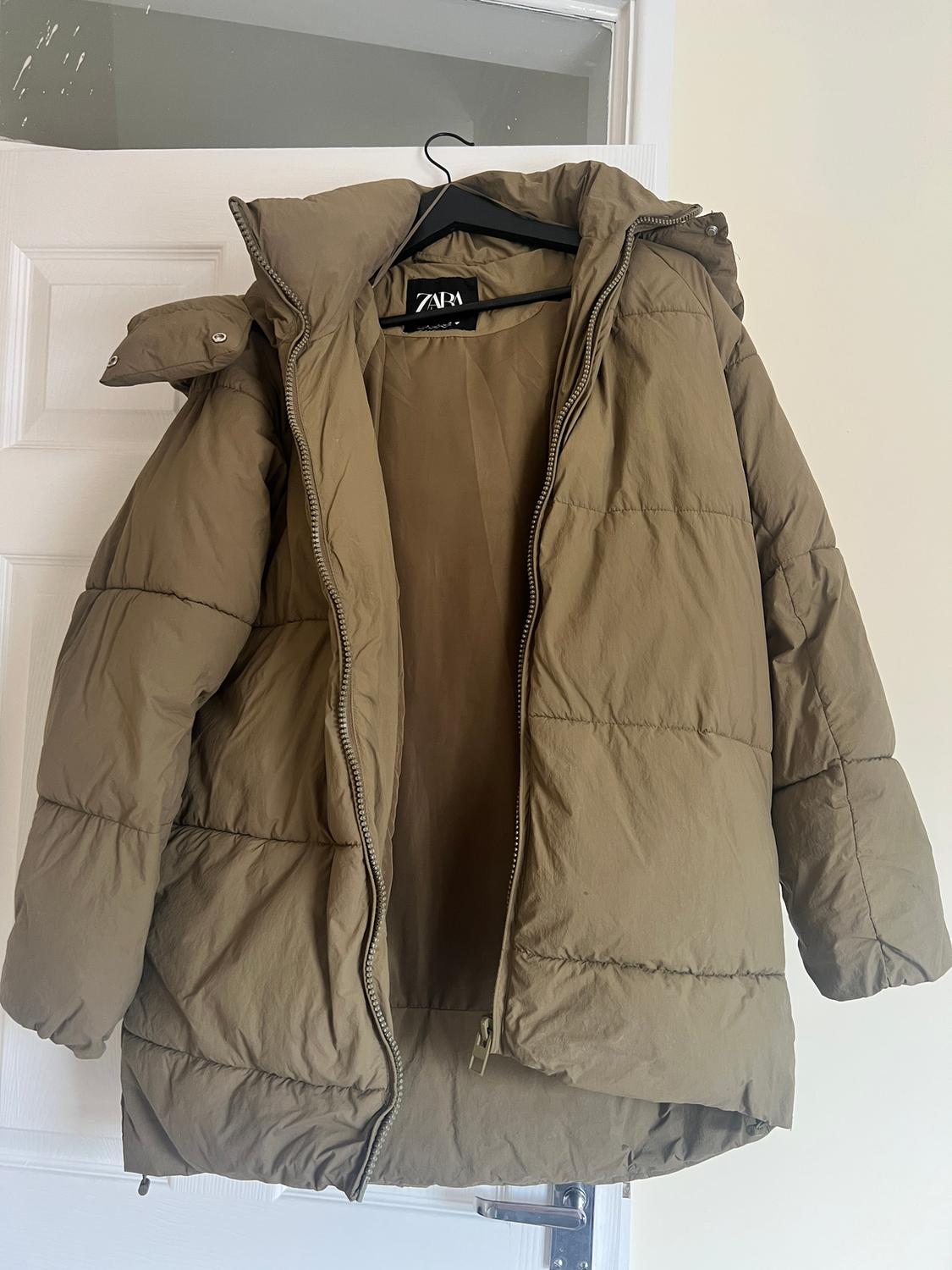 Khaki padded jacket hanging on a black hanger over a white door. Visible label with the text "ZARA".
