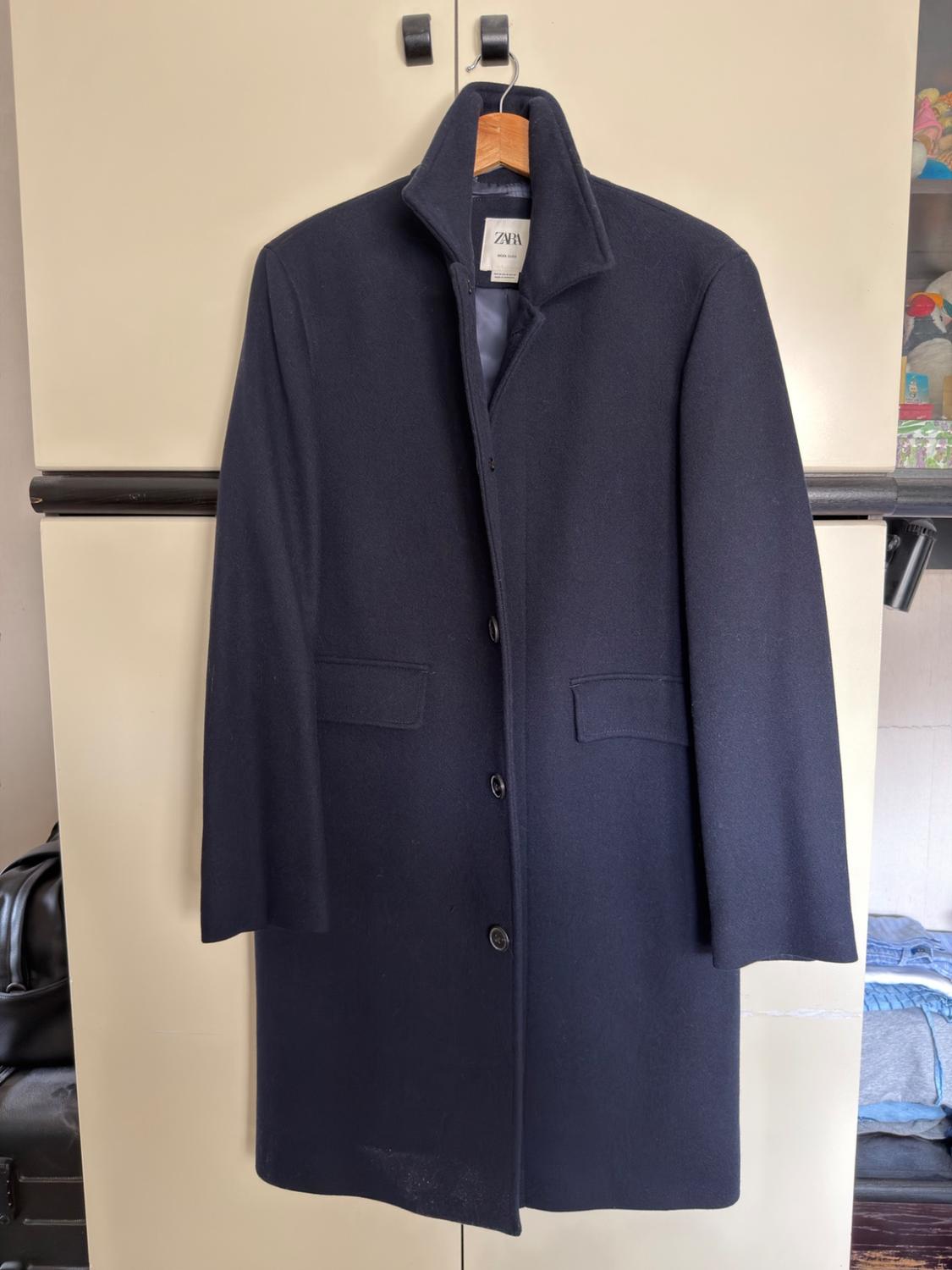Long navy blue coat hanging on a clothes hanger in front of a light-coloured wardrobe. It features buttons, flap pockets and the ZARA label.