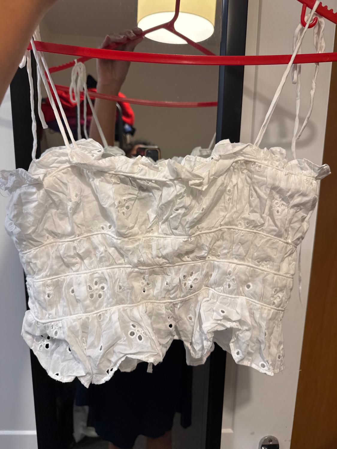 White camisole top with thin straps and floral embroidery, hanging on a red hanger in front of a mirror. Background with clothes and lamp.