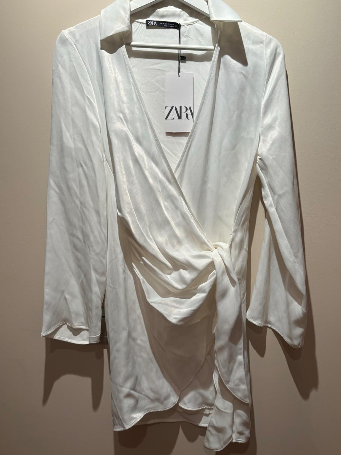 White long-sleeved blouse with collar and waist tie, hanging on a clothes hanger. Label visible with the text "ZARA".