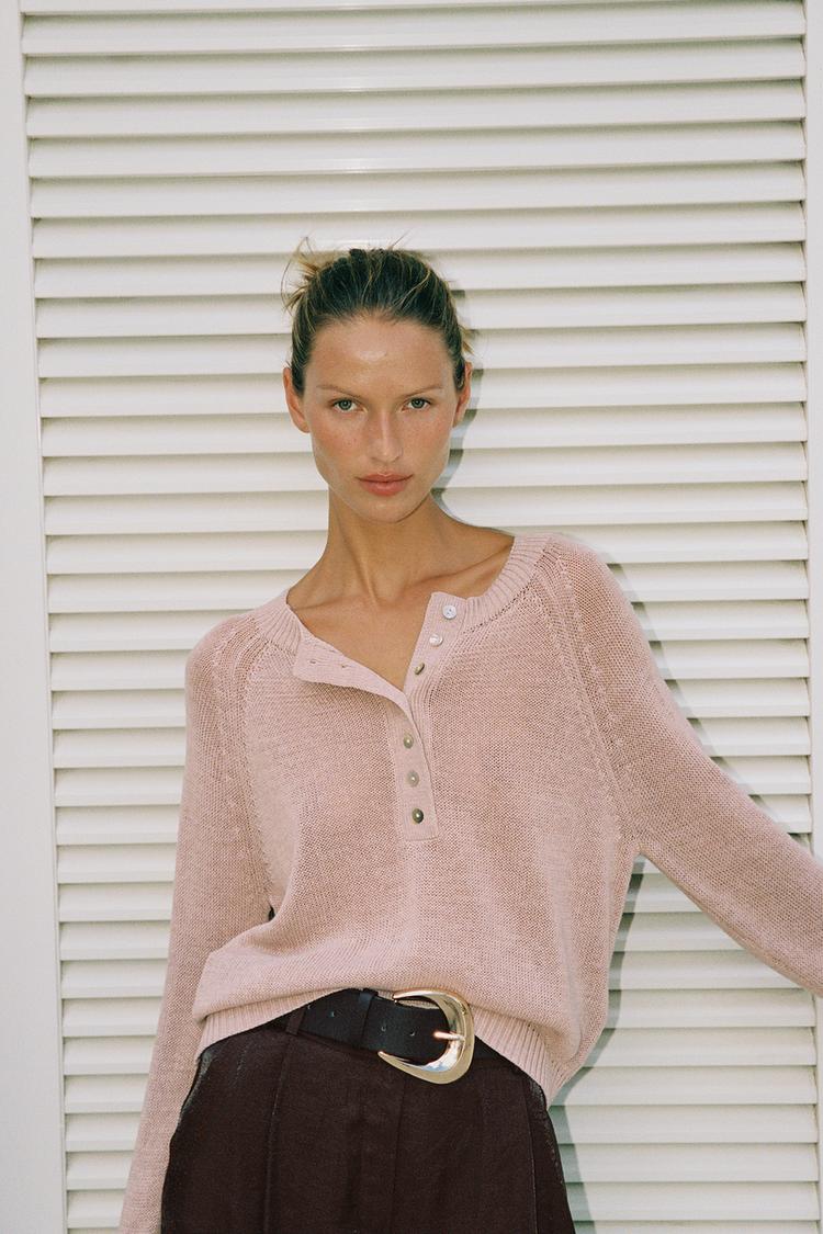 Halbnahe Aufnahme eines lang&auml;rmeligen rosa Pullovers mit Knopfleiste vorne, einer dunklen Hose mit Bundfalten und einem G&uuml;rtel mit Schnalle.