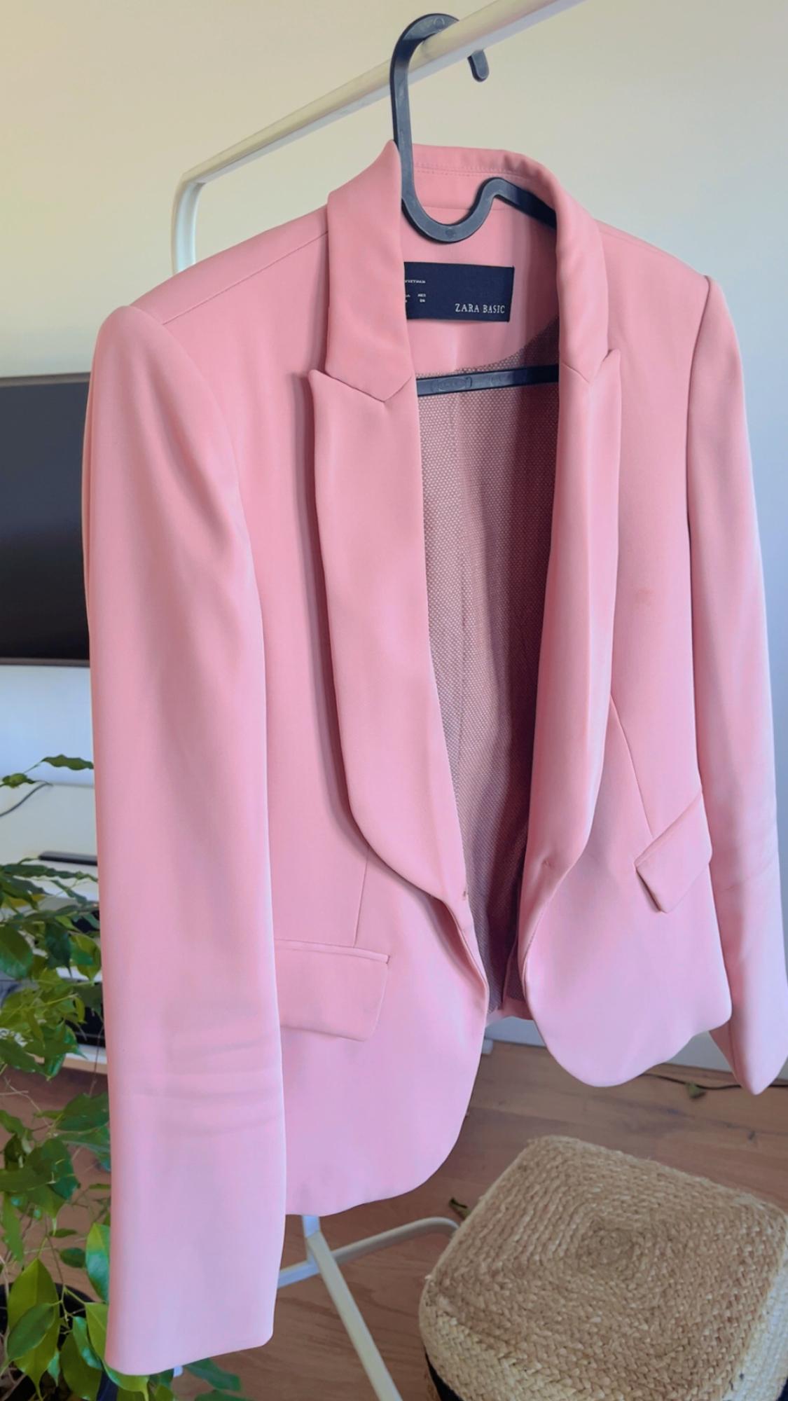 Pink jacket hanging on a black hanger, with a visible label that reads "ZARA BASIC". Background with plant and wicker stool.