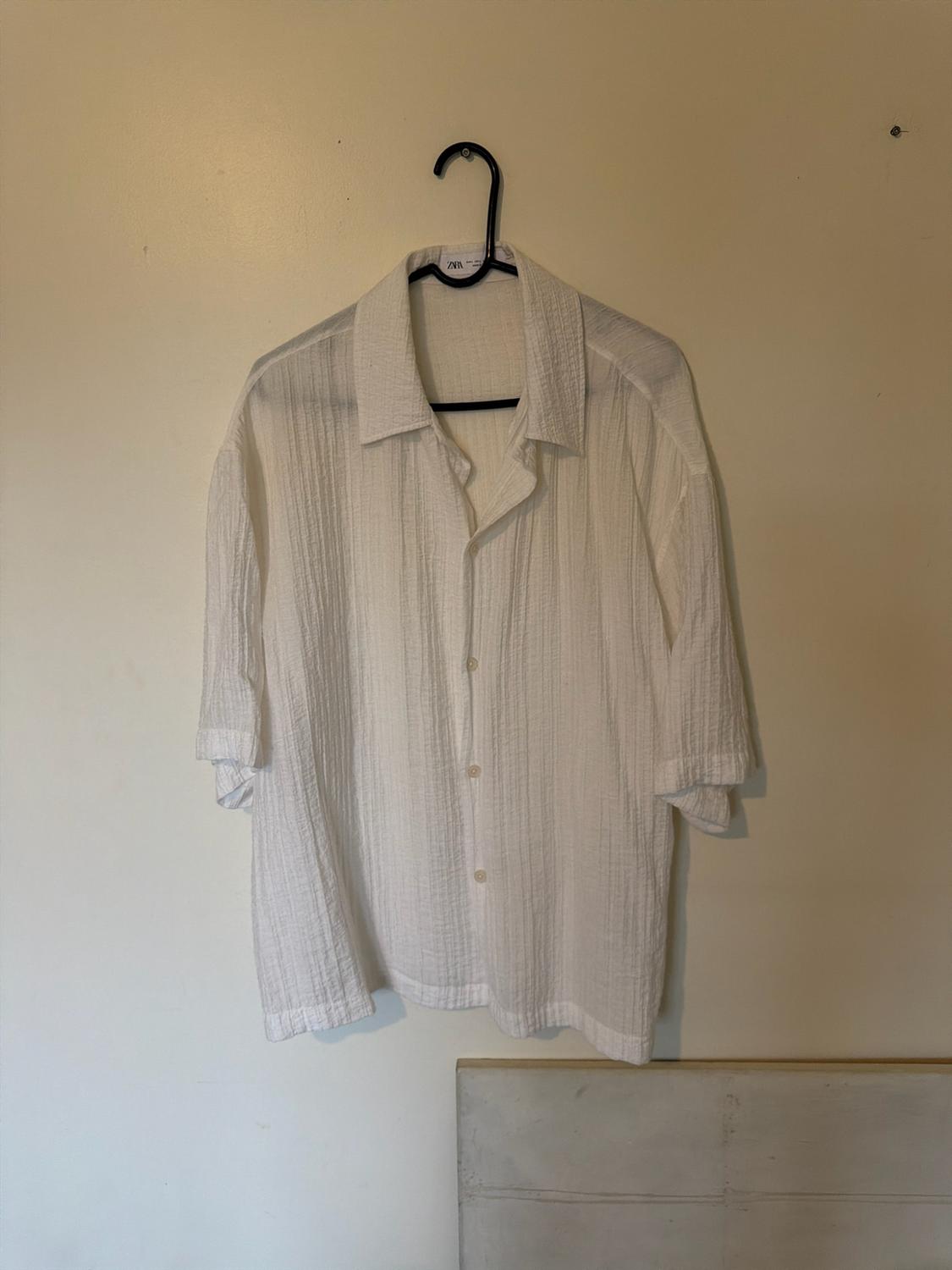 White short-sleeved textured shirt, hanging on a black hanger against a light wall. Relaxed style. Label text: ZARA.