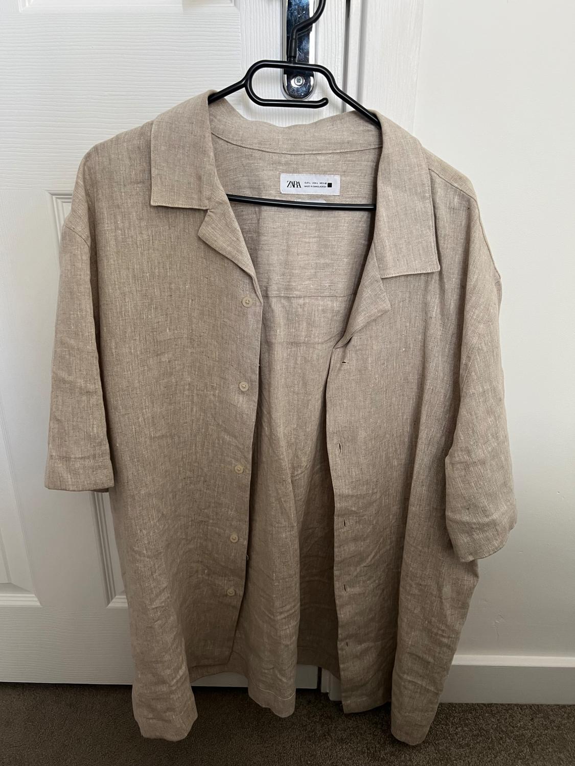 Beige short-sleeve shirt hanging on a hanger from the knob of a white door. Label with text: ZARA EUR L USA L MEX 42.