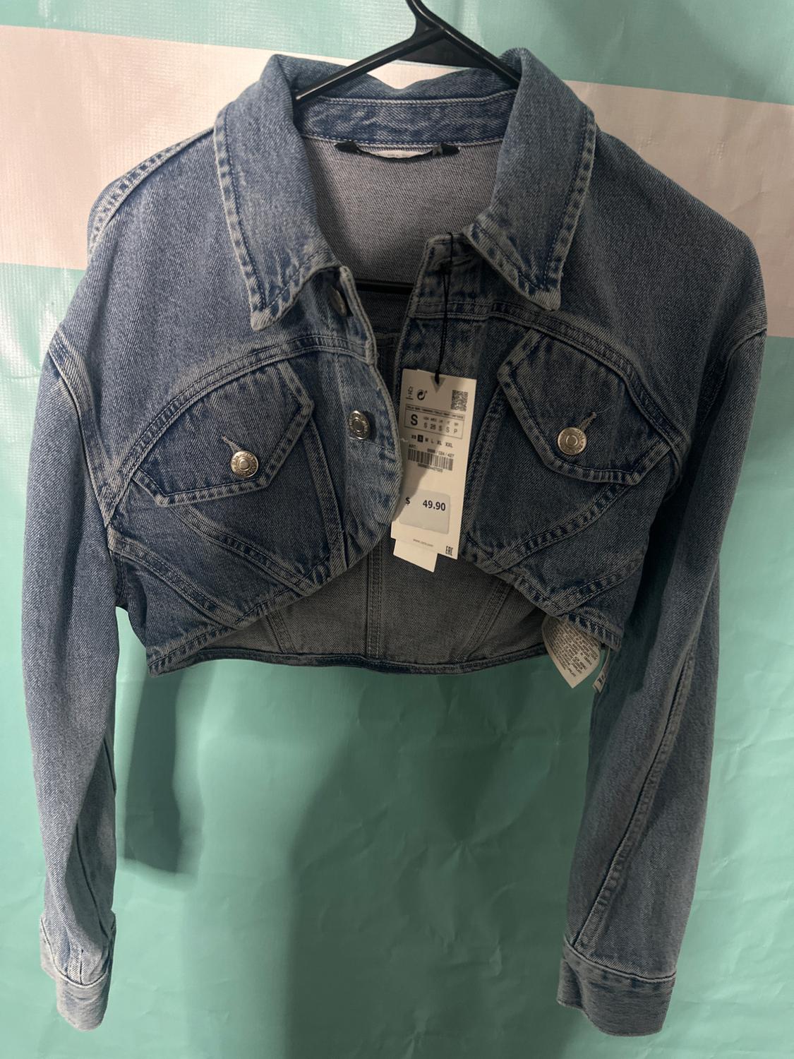 Short blue denim jacket with metal buttons, hanging on a black hanger against a teal background. Visible tag with a price of 49.90.