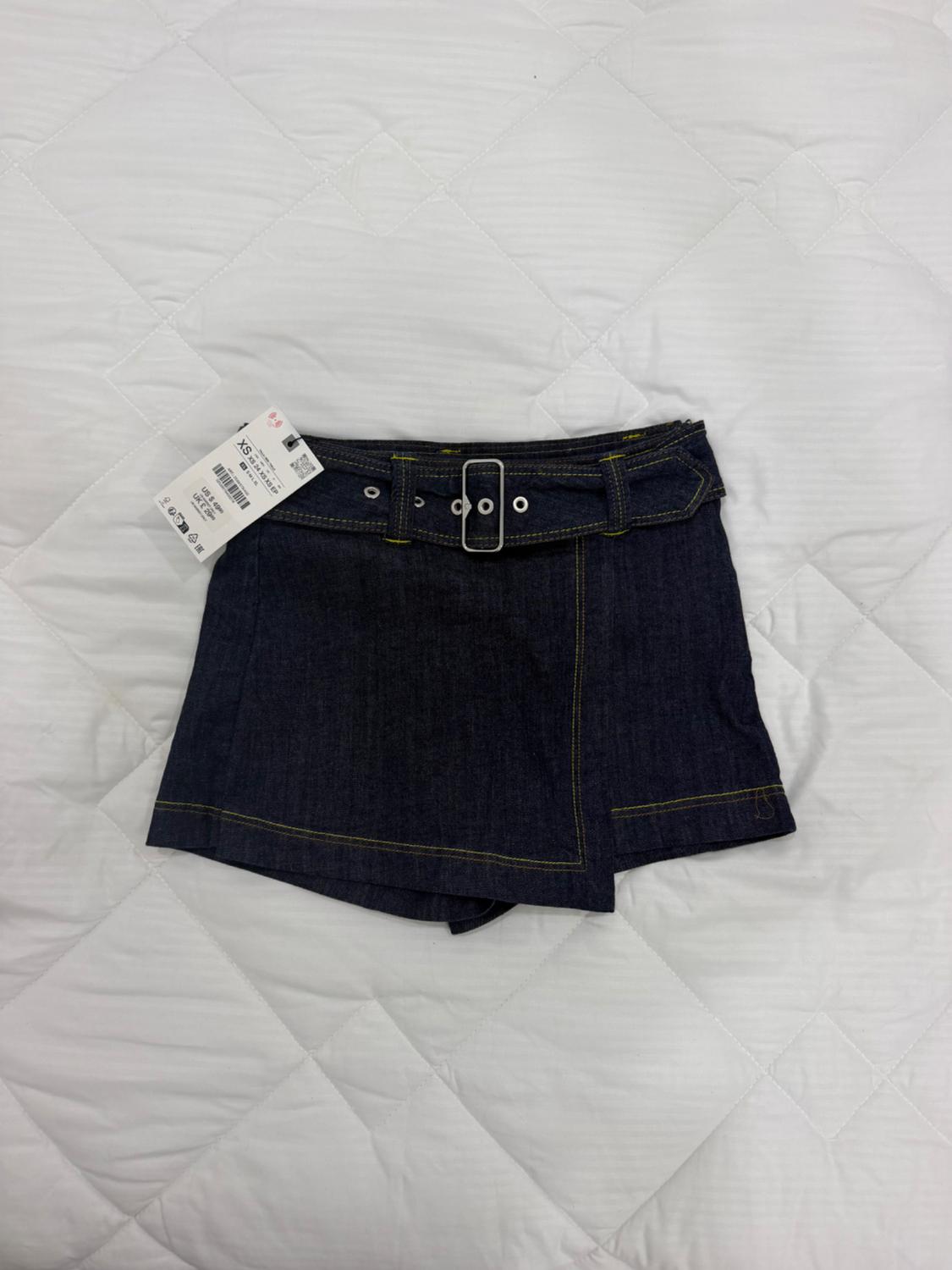 Dark blue asymmetric denim skirt with belt, label, and yellow stitching on a white quilted bedspread.