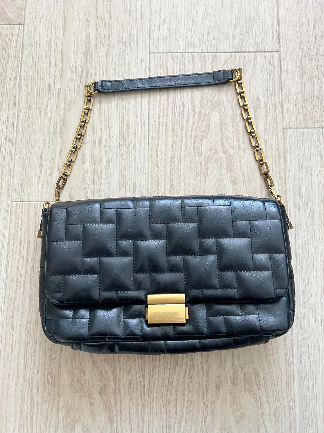 Black quilted handbag with gold clasp and metal chain on a light wooden floor. Elegant and modern design.