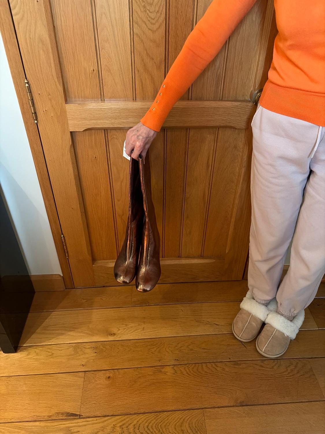 Person in an orange jumper and light-coloured trousers holding brown boots in front of a wooden door. They are wearing slippers with white lining.