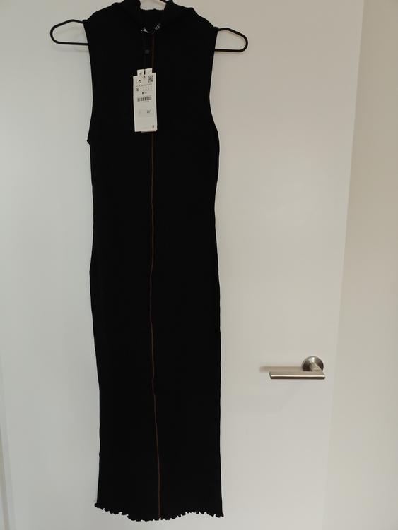 Sleeveless black dress hanging on a hanger over a white door. Visible tag with product details.