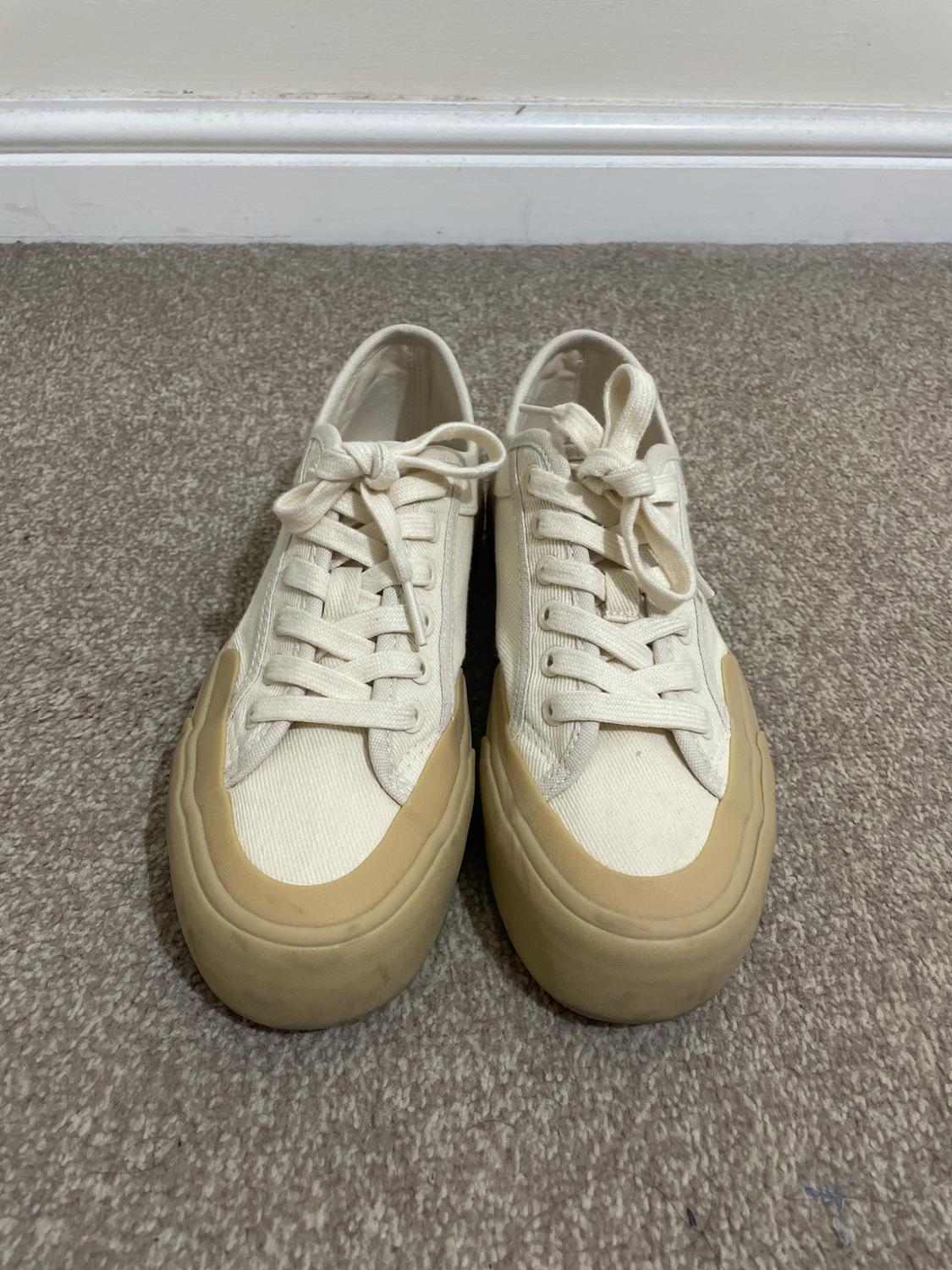 Pair of cream canvas casual trainers with beige rubber soles and toe caps, on a grey carpet next to a white skirting board.