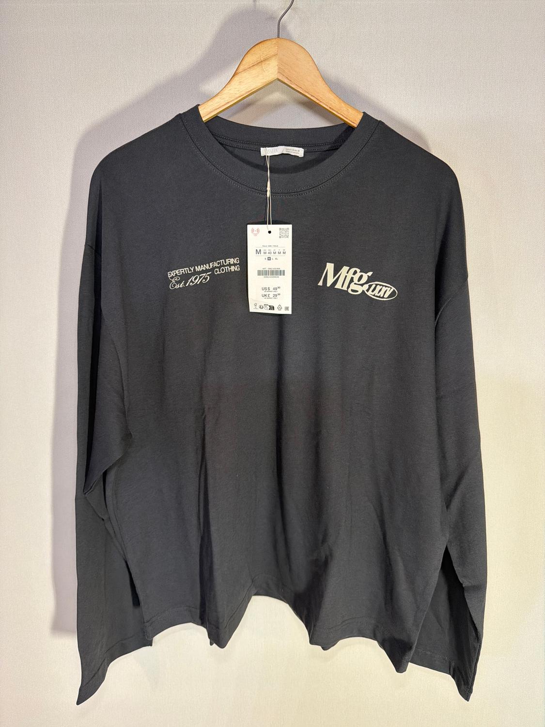 Black long-sleeved t-shirt on a hanger, with text "Expertly Manufacturing Clothing Est. 1975" and "Mfg". Visible hang tag.