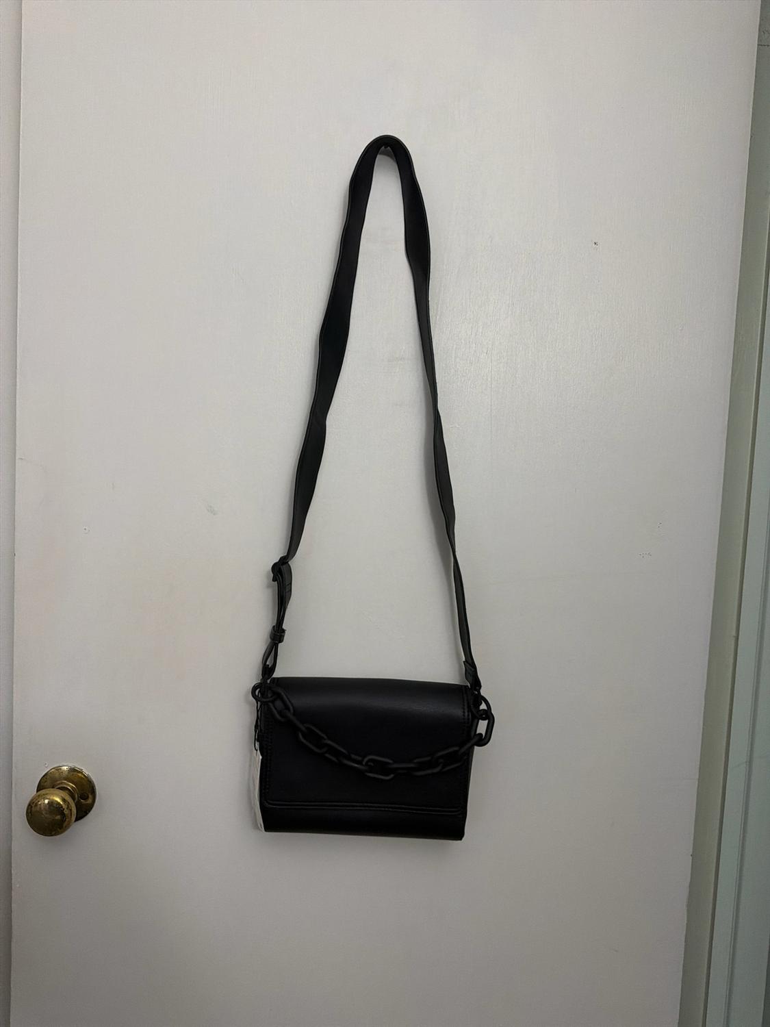 Black bag with a long strap and a chunky decorative chain, hanging on a white door with a gold doorknob.