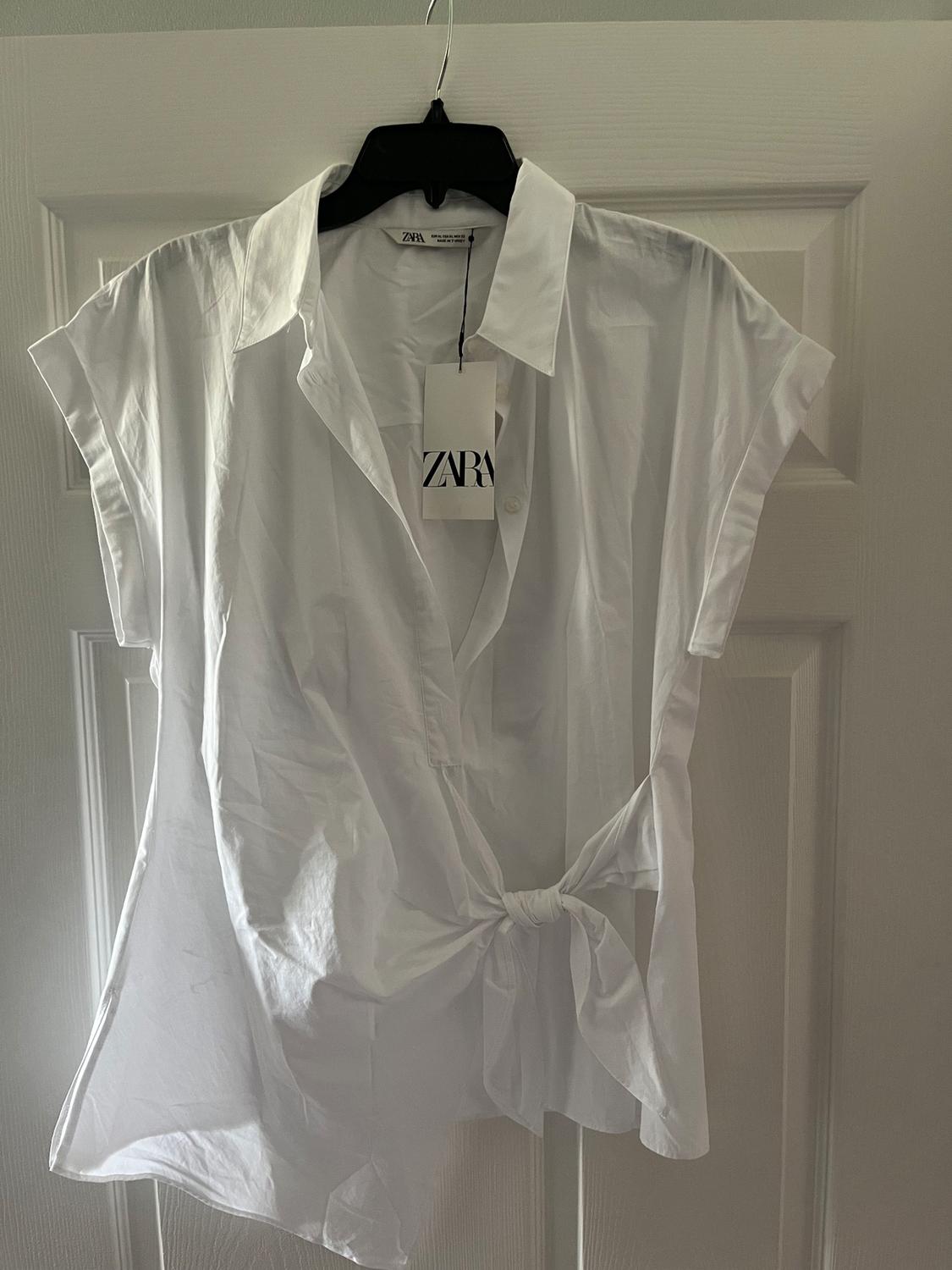 White short-sleeved top with front knot, hanging on a black hanger. Visible label with the text "ZARA".