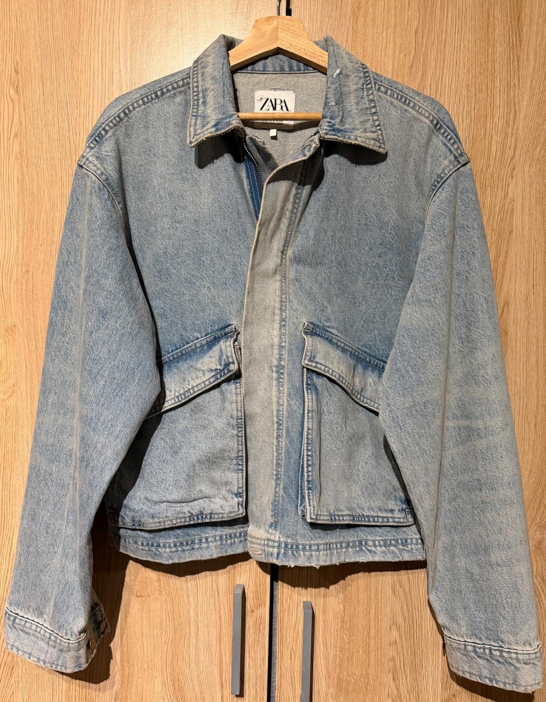 Light blue denim jacket hanging on a wooden hanger, with a visible label that reads "ZARA", against a wooden background.
