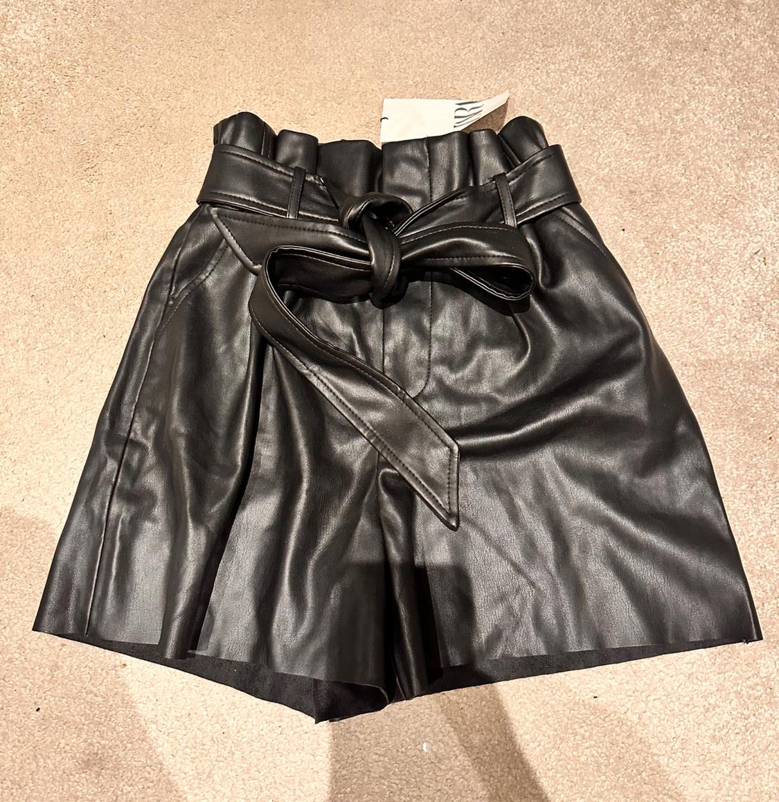 Black faux leather shorts with tie belt, on beige background.