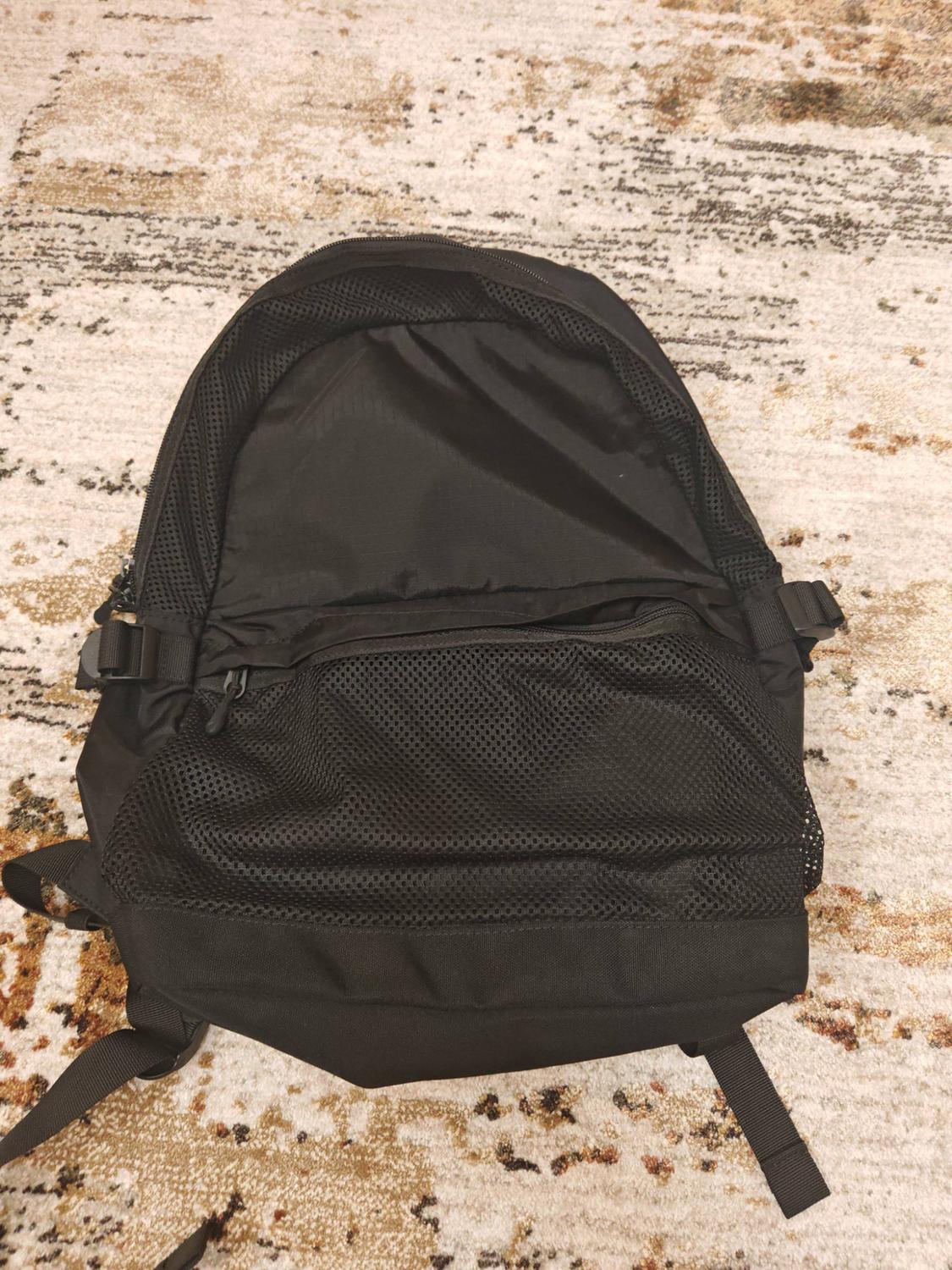 Black backpack with a front mesh pocket, viewed from above on a beige patterned rug. Casual and relaxed atmosphere.