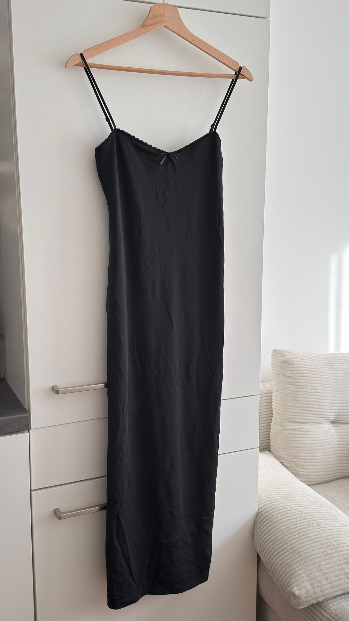 Black spaghetti-strap dress hanging on a wooden hanger over a white wardrobe, next to a light-coloured sofa.