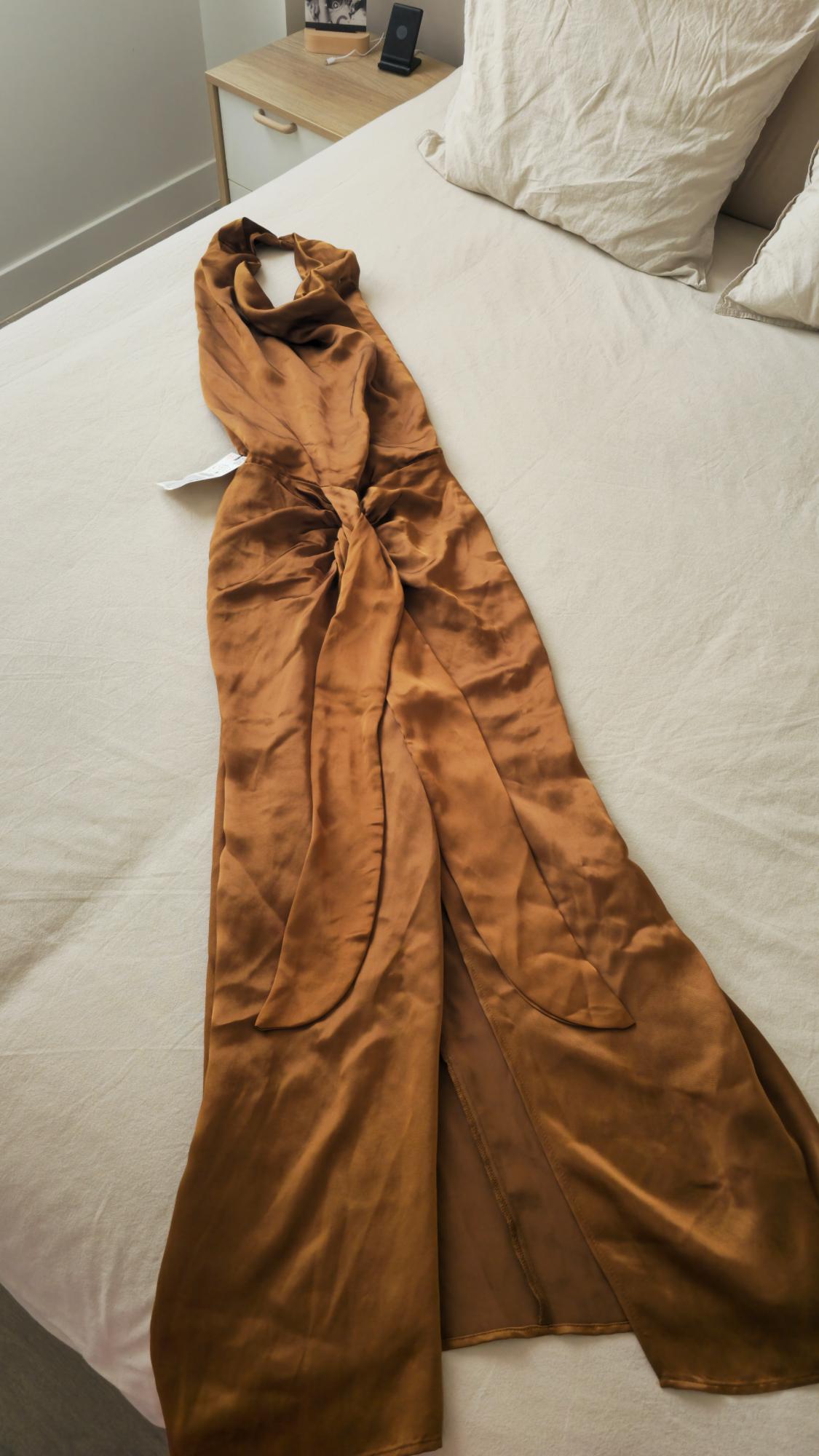 Brown satin dress spread out on a bed with light sheets and pillows, next to a wooden bedside table.