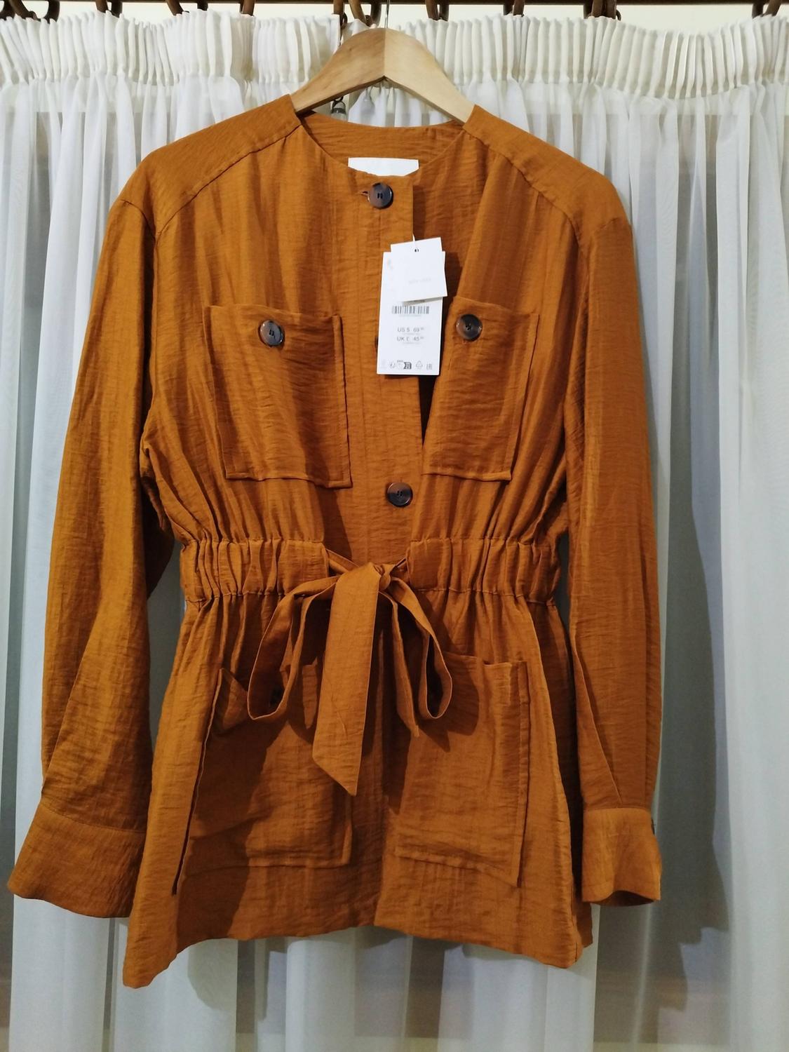 Brown jacket with buttons, pockets, a belt and a label, hanging on a wooden hanger in front of white curtains.