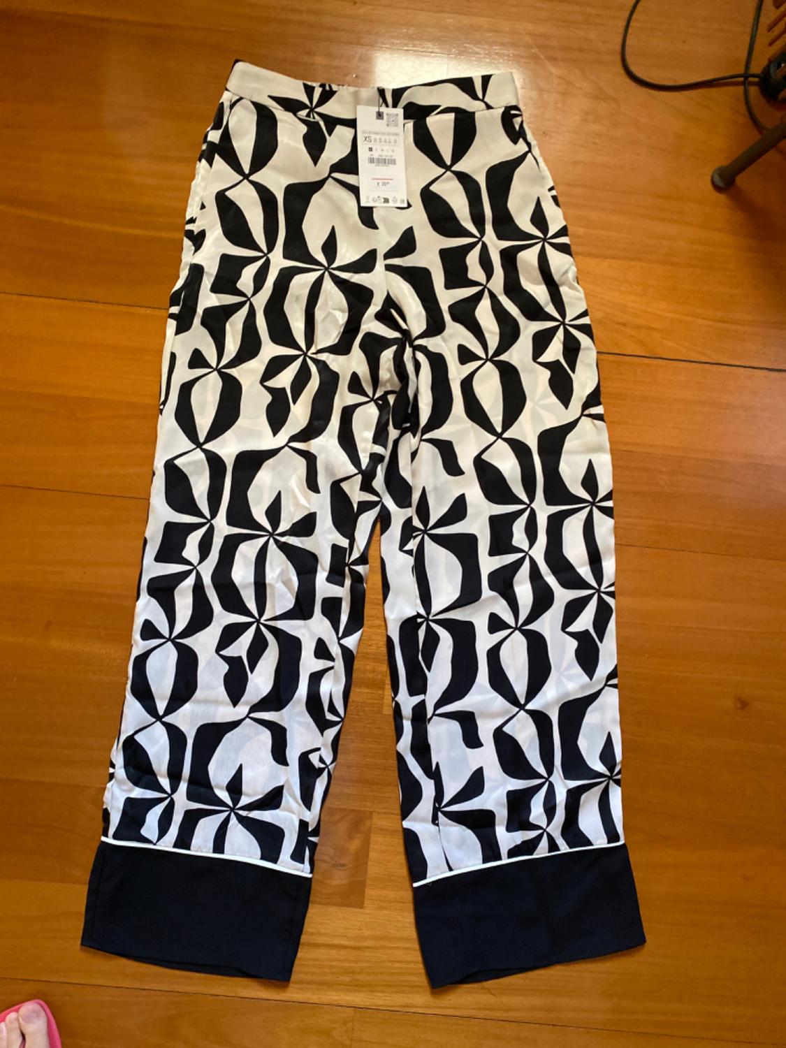 White trousers with a black geometric print and dark hem, on a wooden floor. Label visible at the waist.