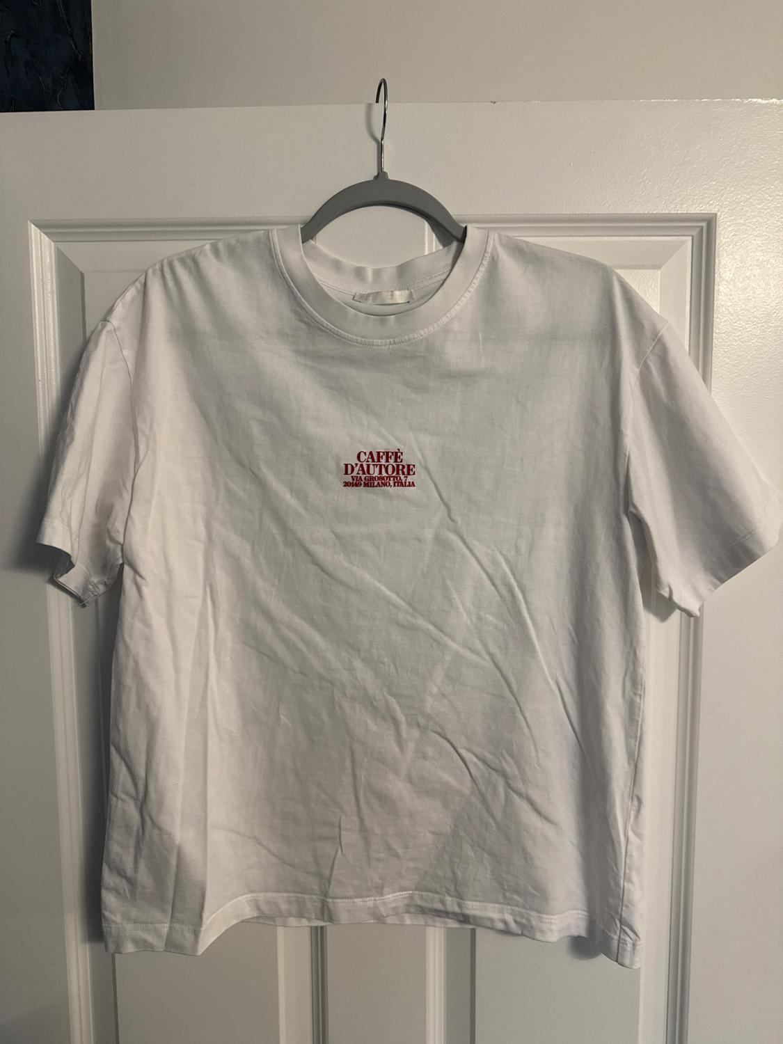 White t-shirt hanging on a hanger, with red text "Caffè D'Autore 2016 Milano Italia" on the chest, over a white door.