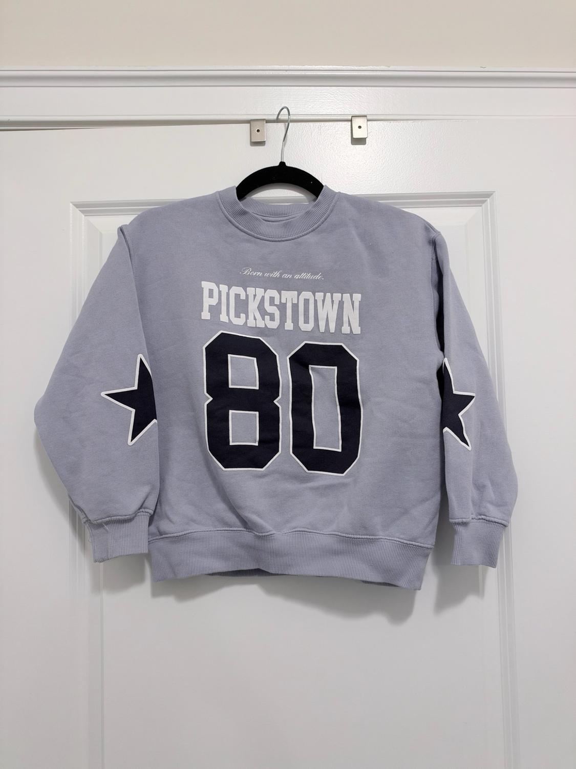 Light blue sweatshirt hanging on a white door. Texts: "Born with an attitude.", "PICKSTOWN", "80". Star detail on the sleeves.