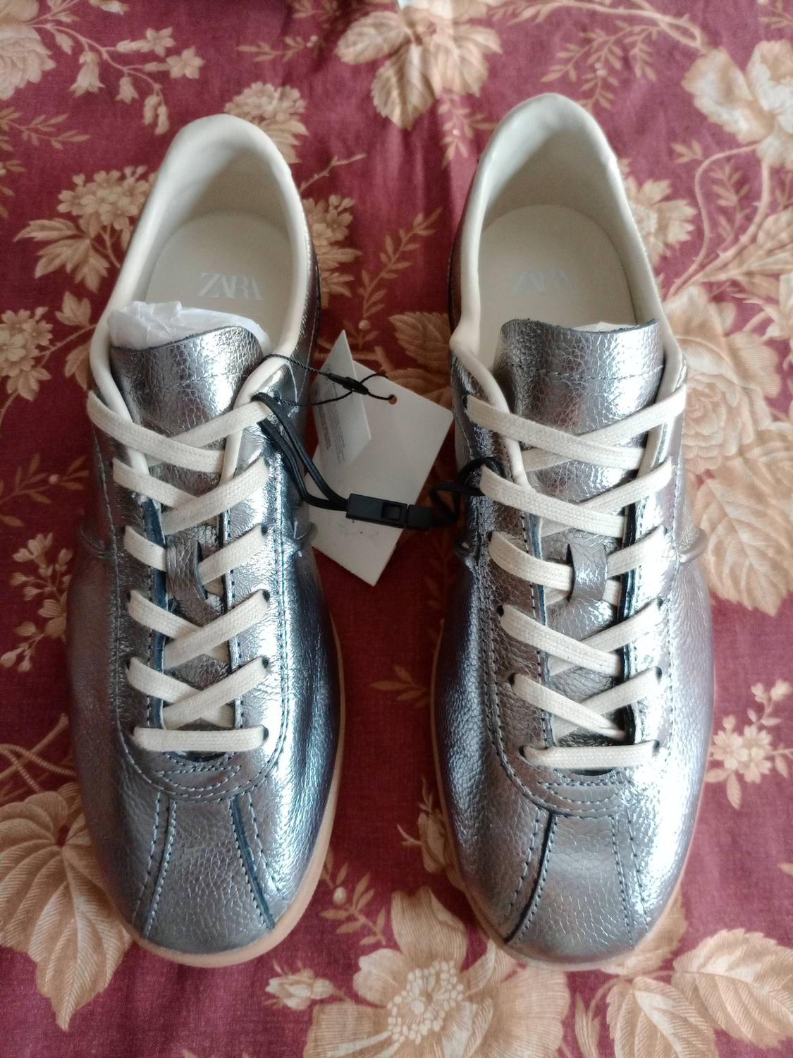 Metallic silver trainers with white laces on red floral print fabric. ZARA text on the inside.