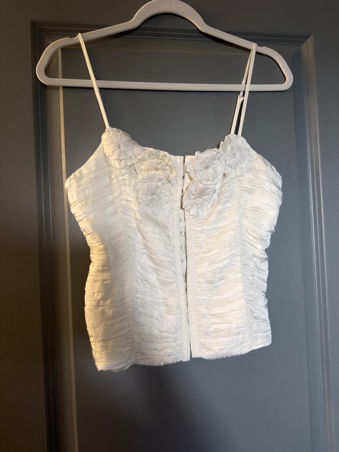 White strappy top with ruched fabric, flowers on the neckline and hook-and-eye fastening, hanging on a white hanger in front of a grey door.