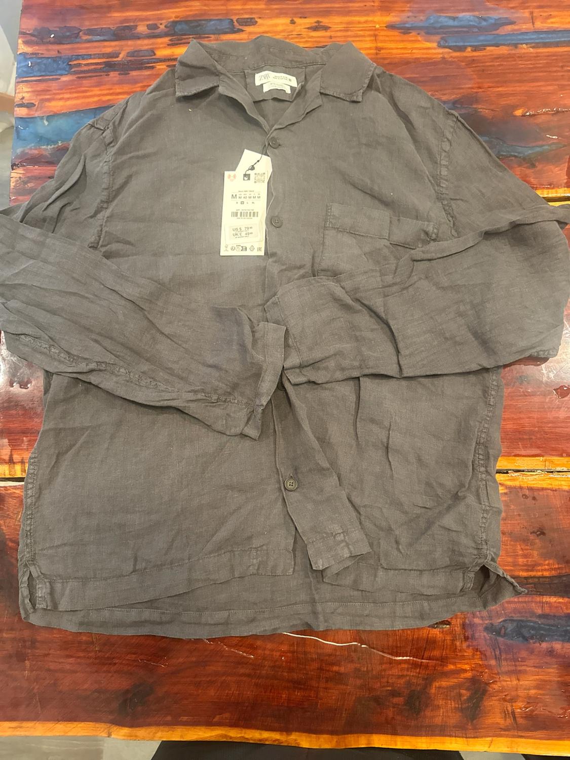 Dark grey button-down shirt with a pocket and ZARA label, folded on a rustic wooden table with reddish and bluish tones.