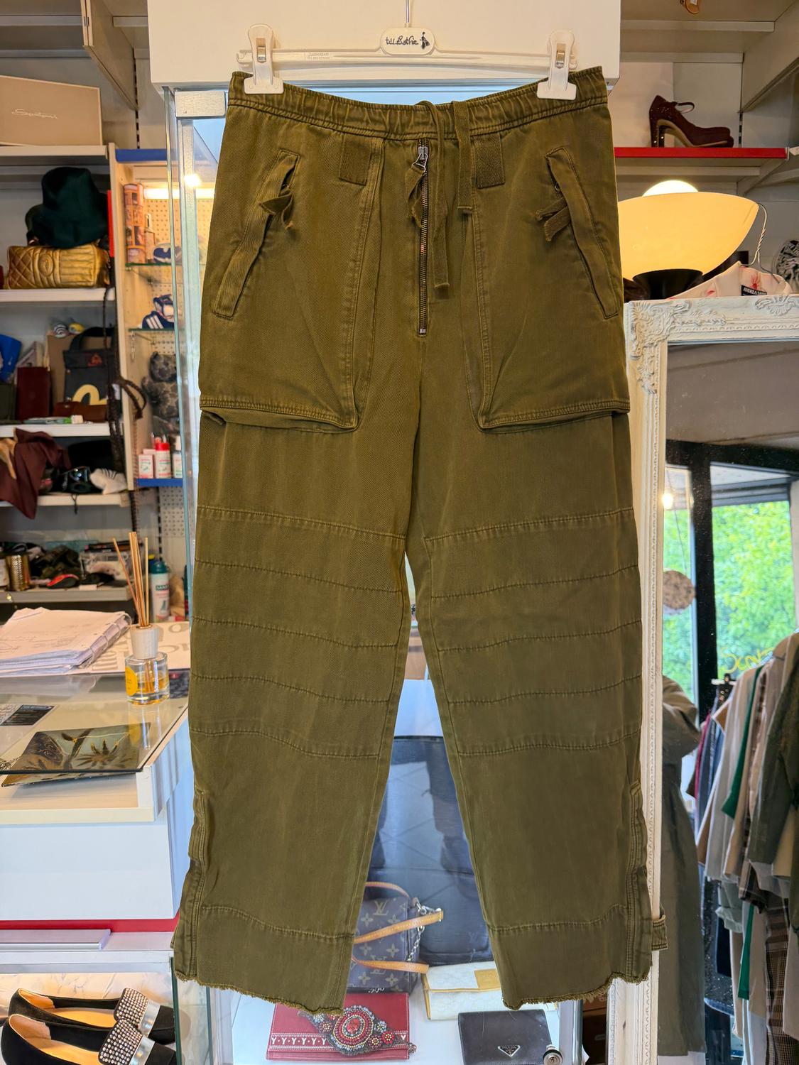 Green cargo trousers hanging on a hanger in a shop, with large pockets and an adjustable drawstring. Background with shelves and accessories.