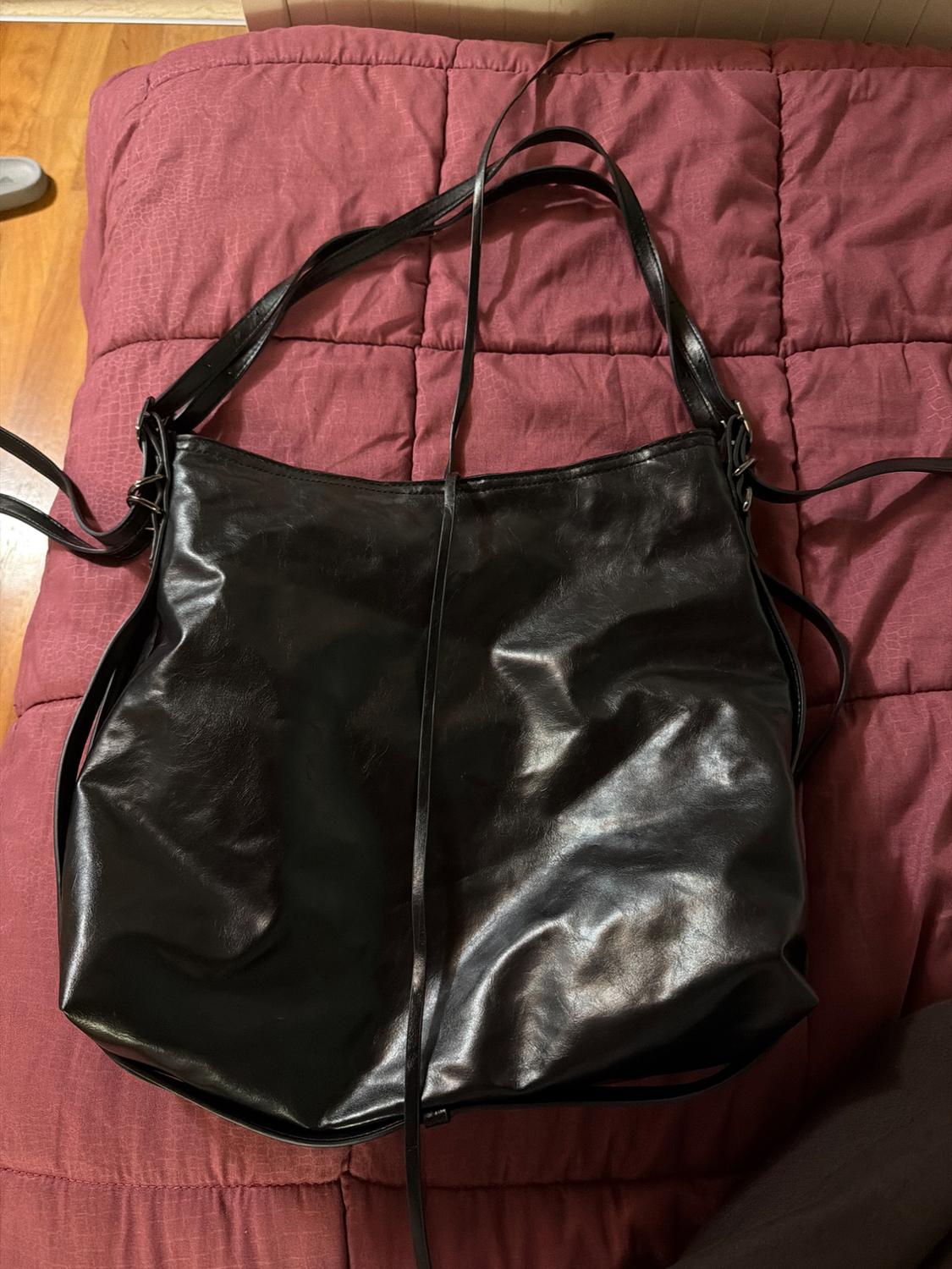 Large black leather handbag with long straps, on a red quilted bedspread. Light wood background.