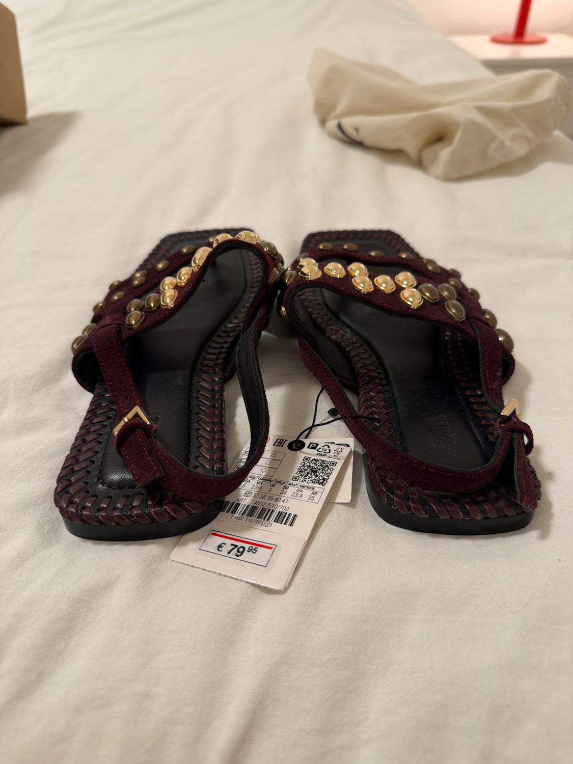 Dark brown leather sandals with gold studs on a white bed. Price tag visible: €79.95.