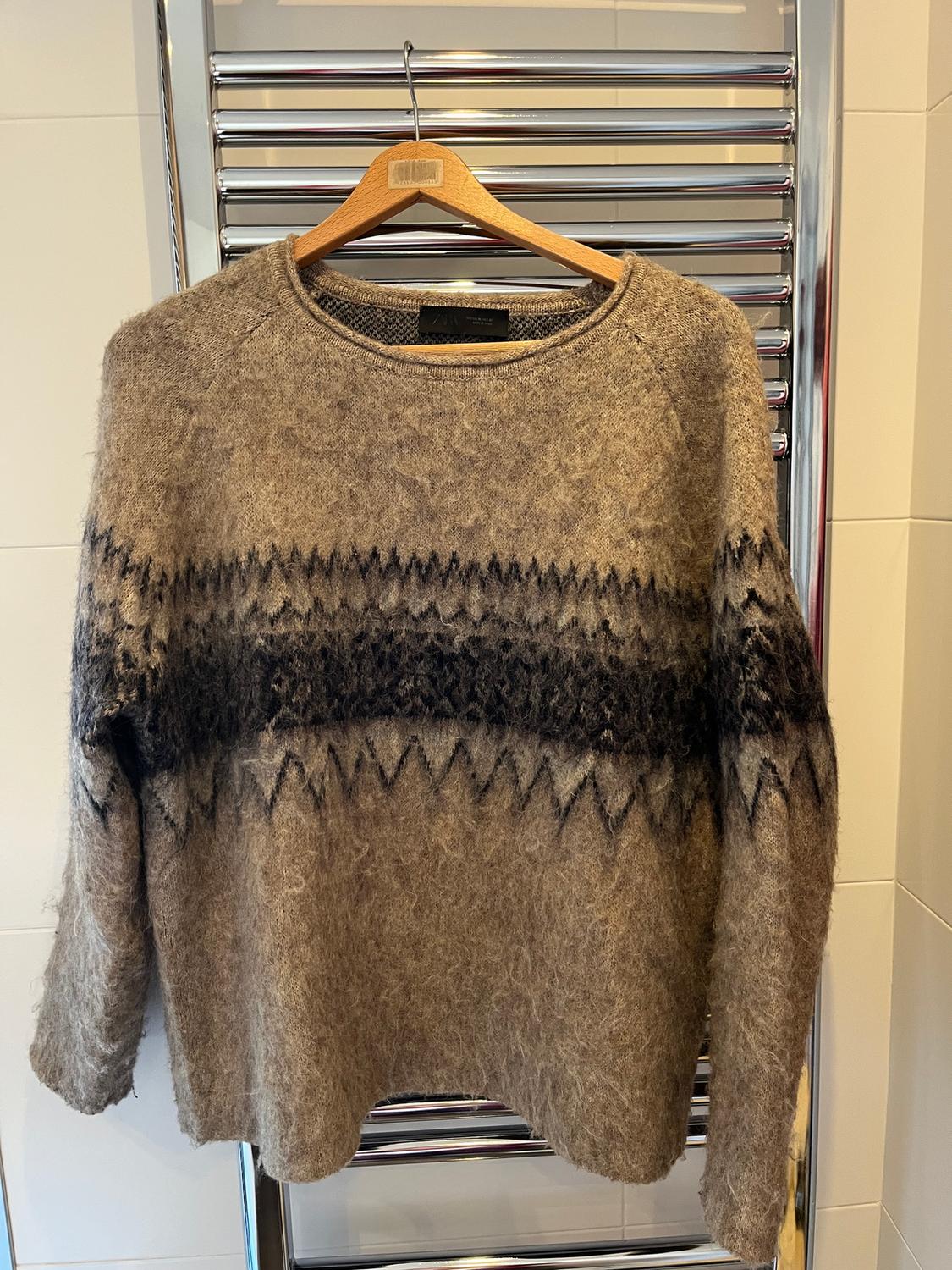 Brown wool jumper with black geometric pattern hanging on a wooden hanger over a chrome radiator.
