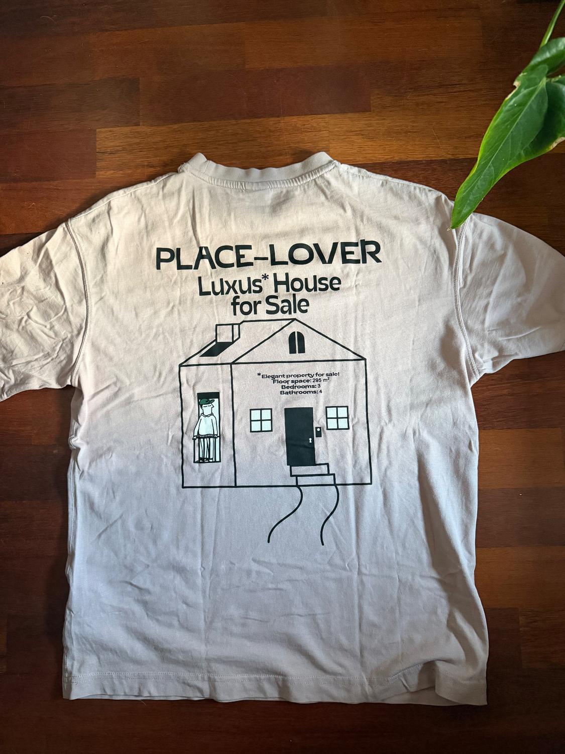 Λευκό t-shirt σε ξύλινο πάτωμα με σχέδιο σπιτιού και κείμενο: "PLACE-LOVER Luxus* House for Sale". Φυτό και πράσινα φύλλα ορατά.