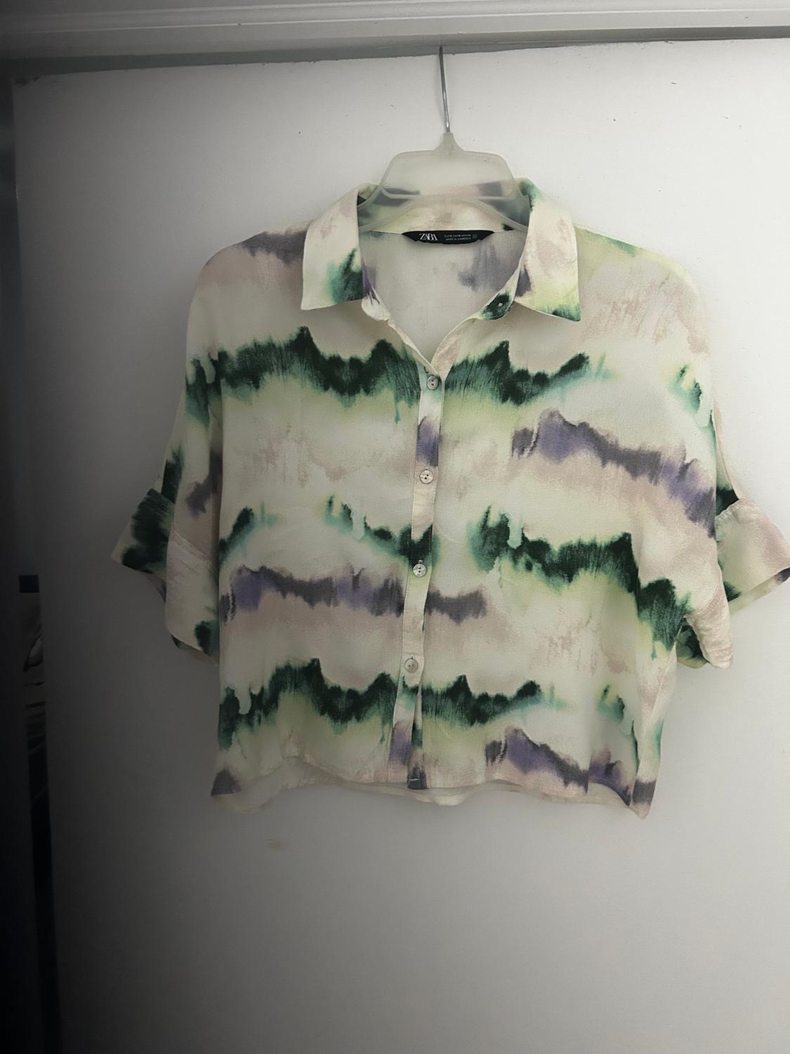Short shirt with abstract green and purple print, hanging on a clear hanger against a white background.
