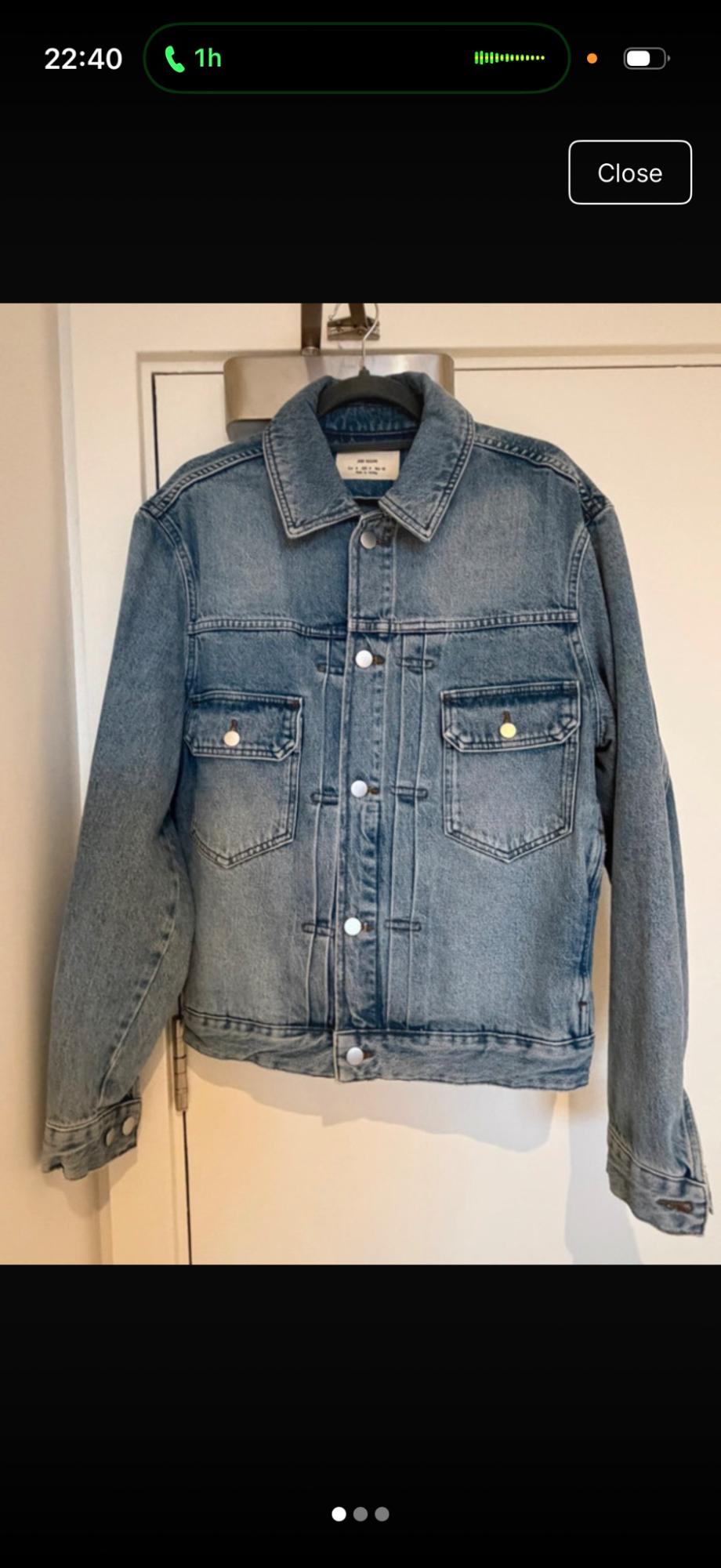 Blue denim jacket hanging on a hanger over a white door. It features a silver button fastening and two chest pockets.