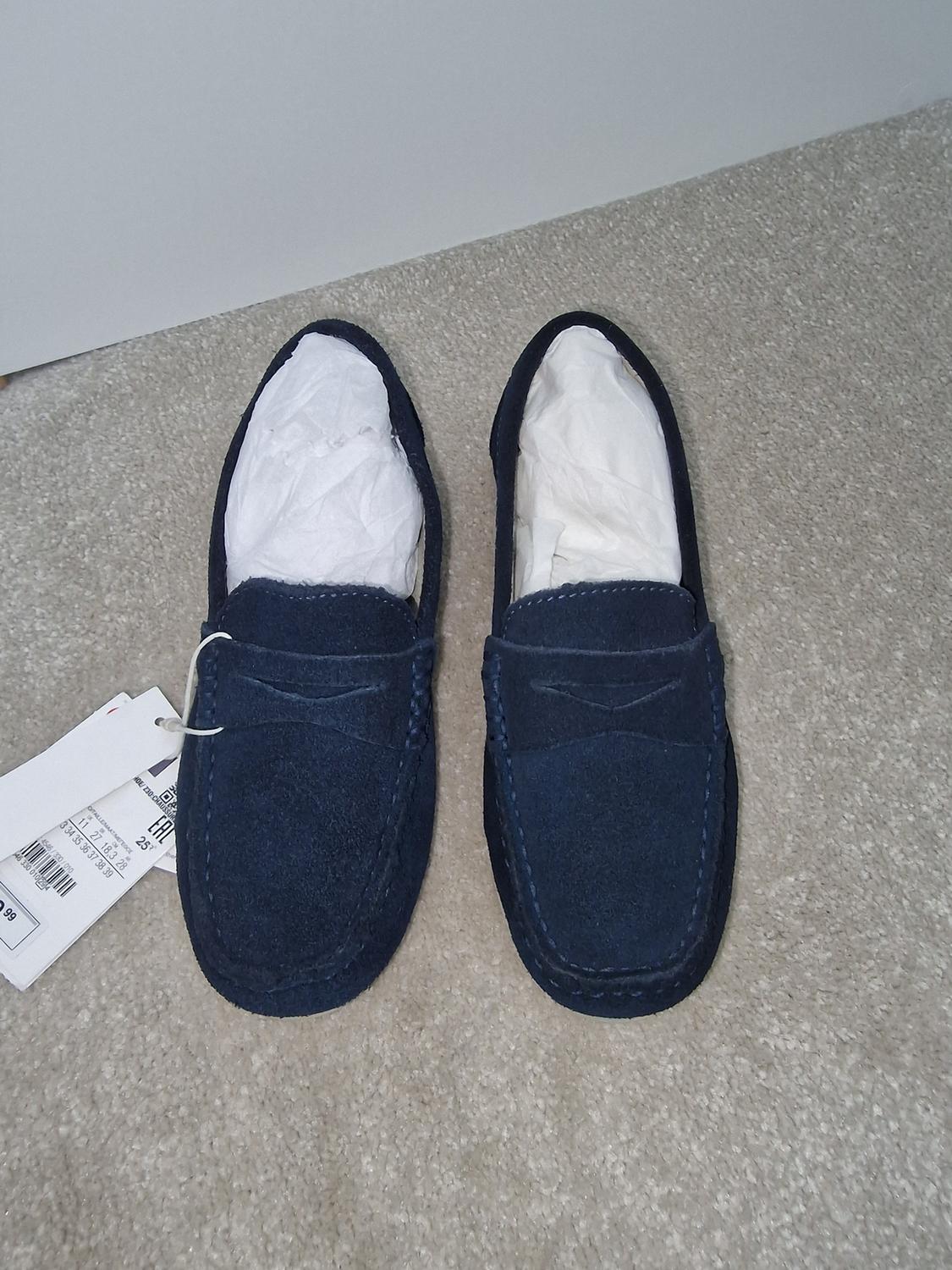Dark blue suede shoes on a beige carpet, with price tags visible on the left.