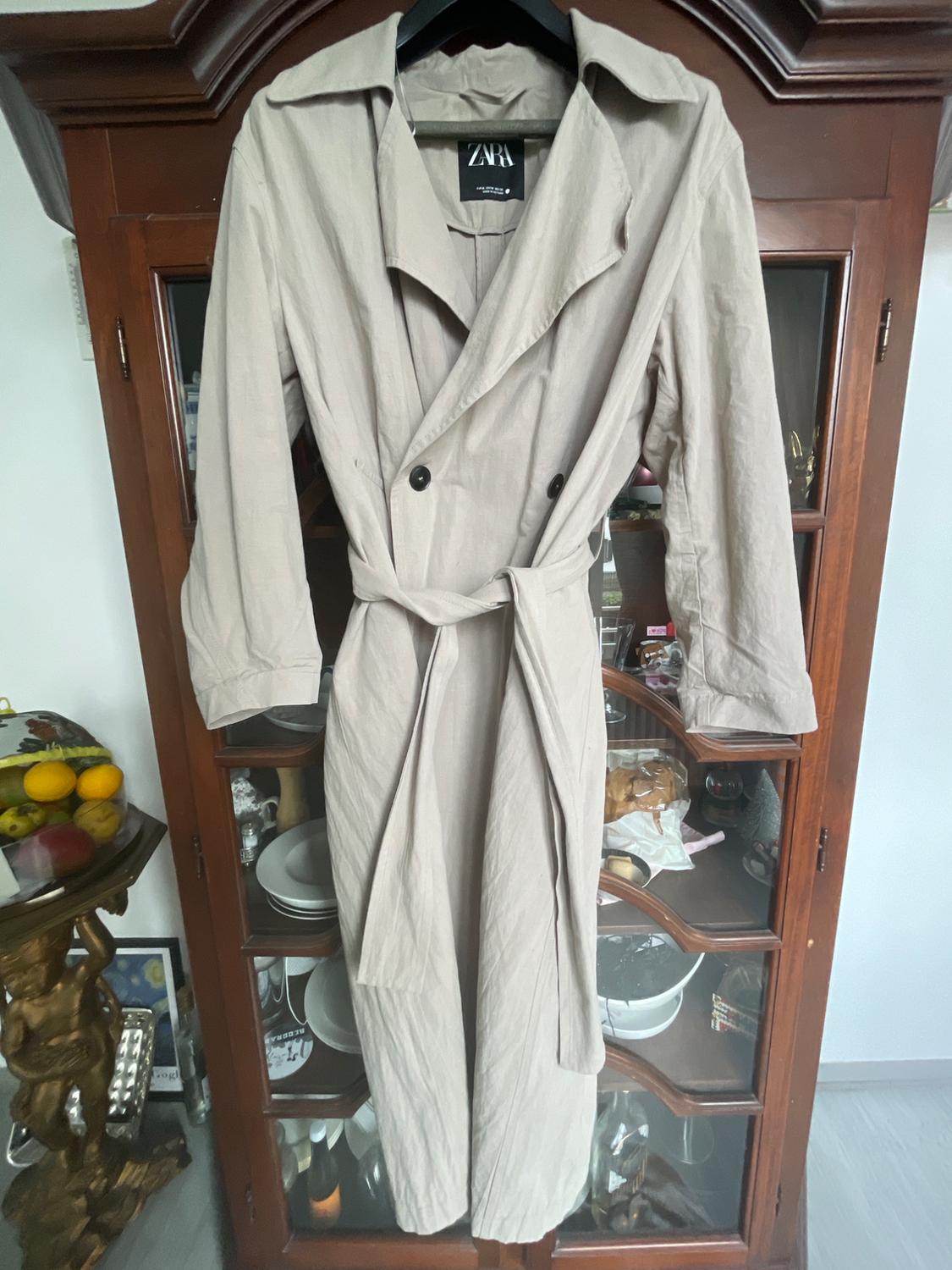 Beige trench coat hanging on a hanger in front of a wooden wardrobe with glass doors. Visible label: ZARA.