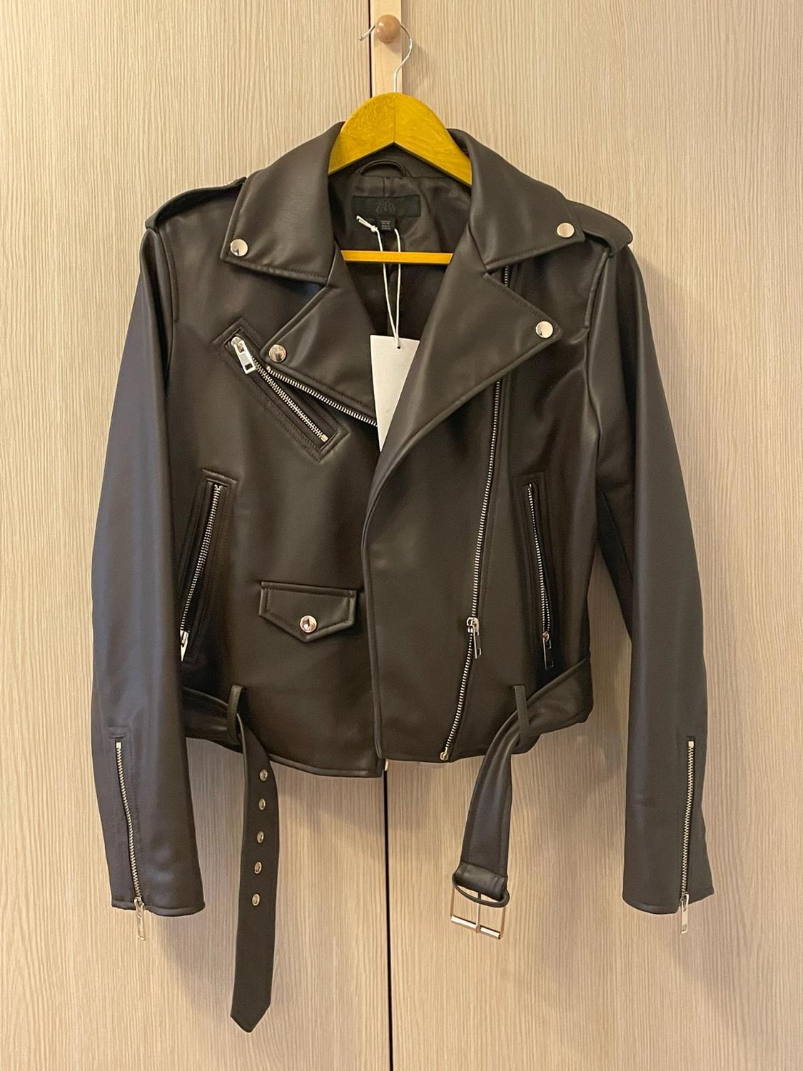 Black biker-style jacket with zips and a belt, hanging on a yellow coat hanger against a light wood background.