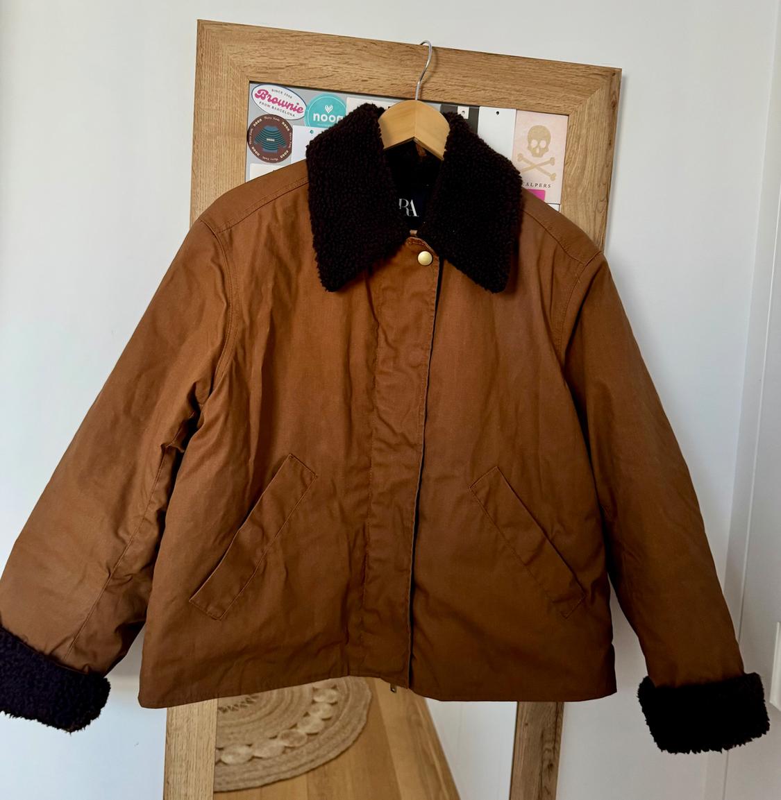 Brown jacket with black wool collar and cuffs hanging on a wooden coat rack. Background with colourful stickers.
