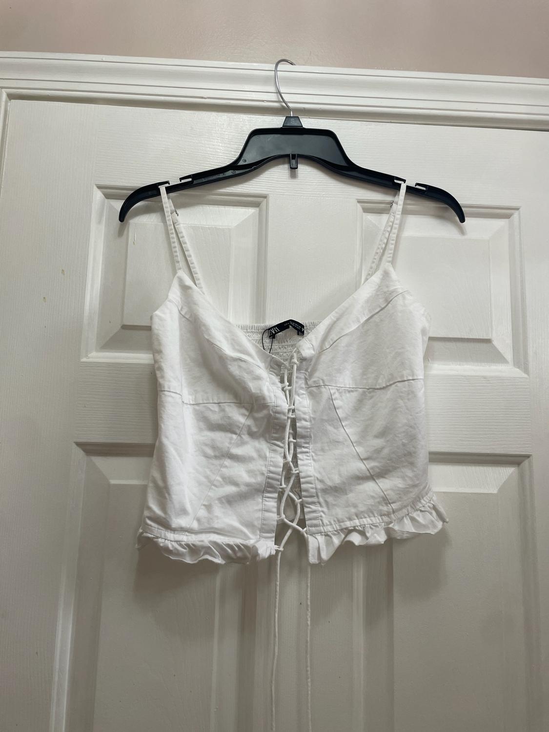 White strappy top with front lacing and a ruffled hem, hanging on a hanger over a white door.