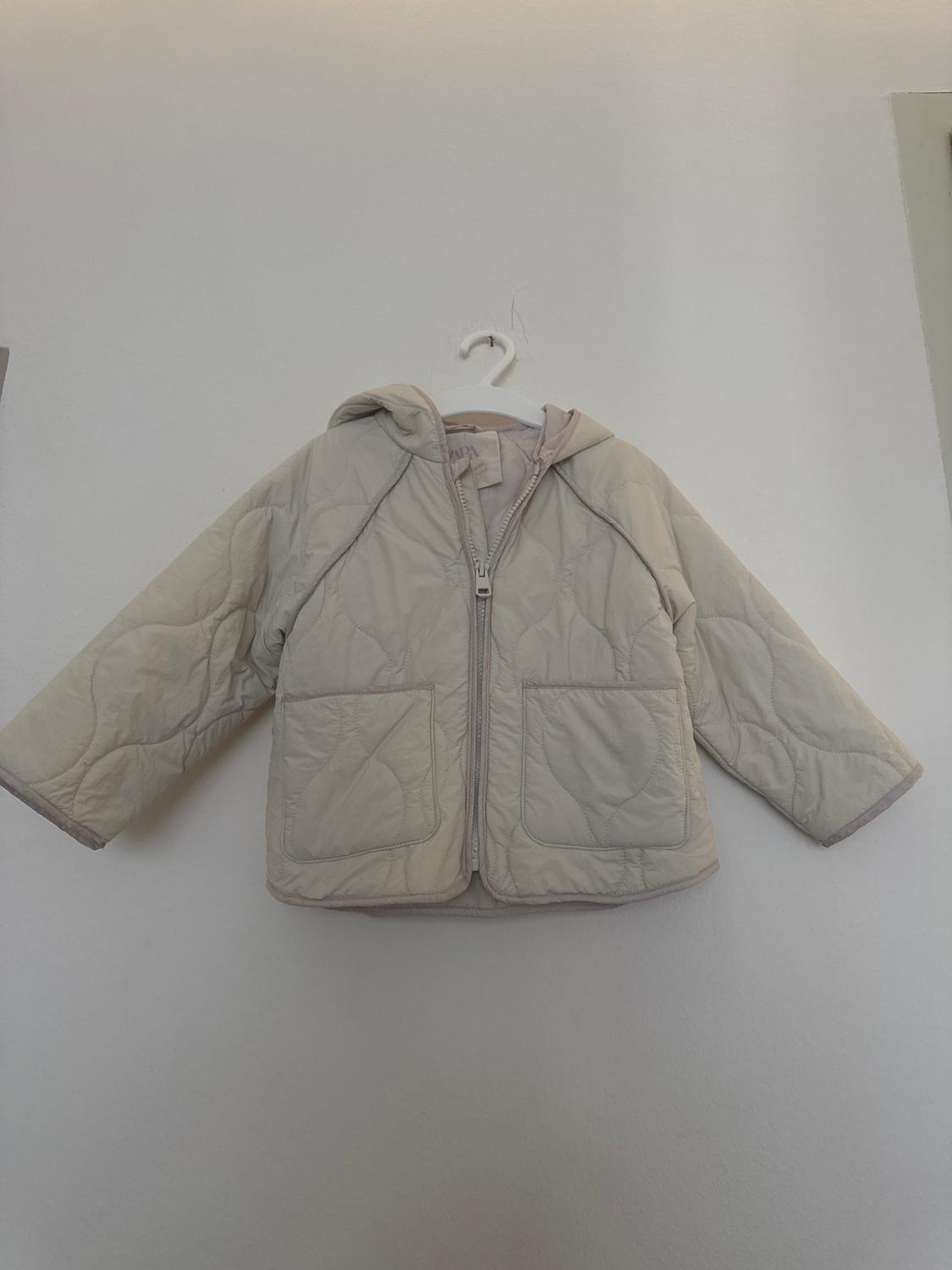 Beige padded jacket with hood and zip, hanging on a white hanger against a light-coloured wall.